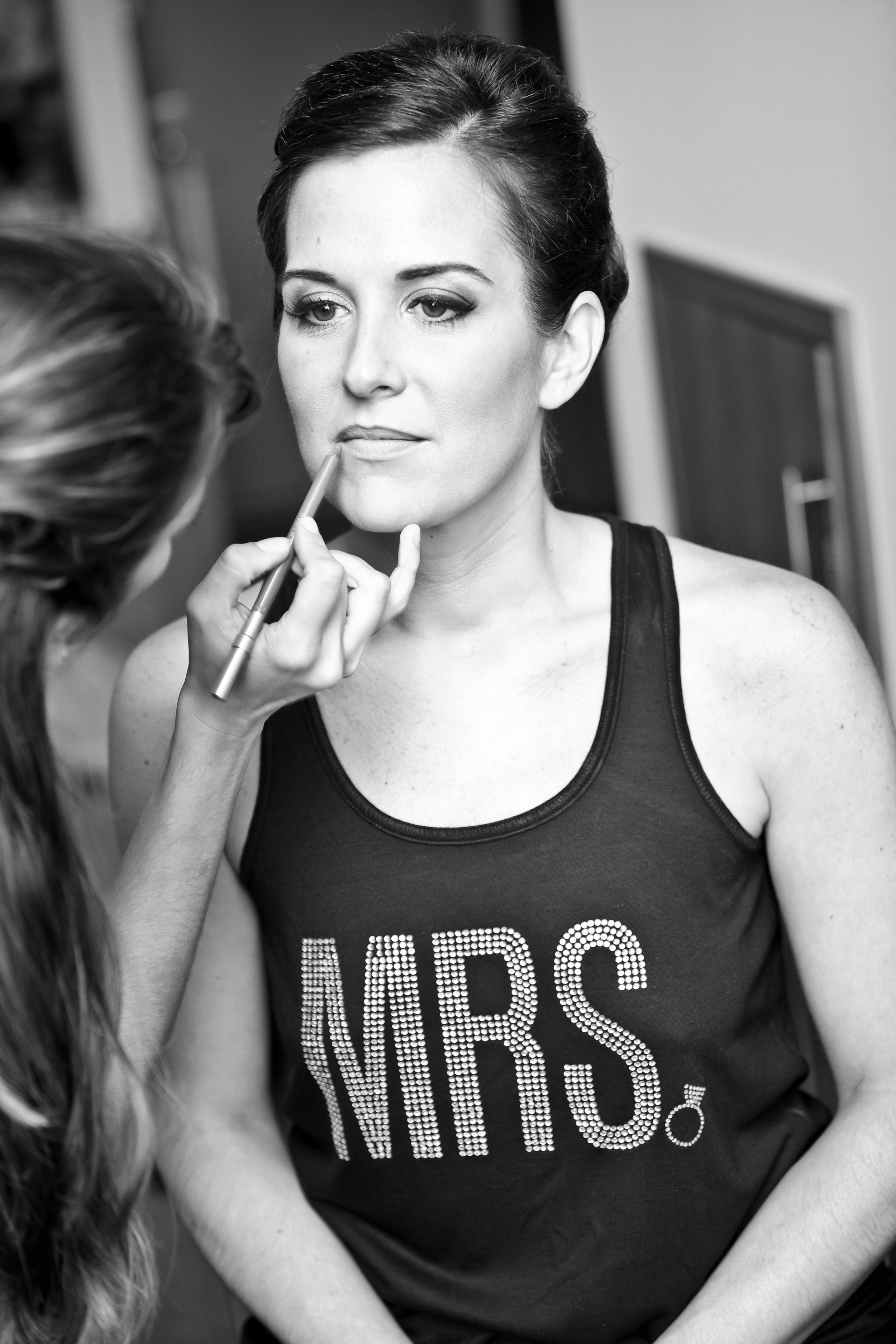 Woman with dark hair styled in an updo getting makeup applied by another person, wearing a tank top that says 'MRS.'