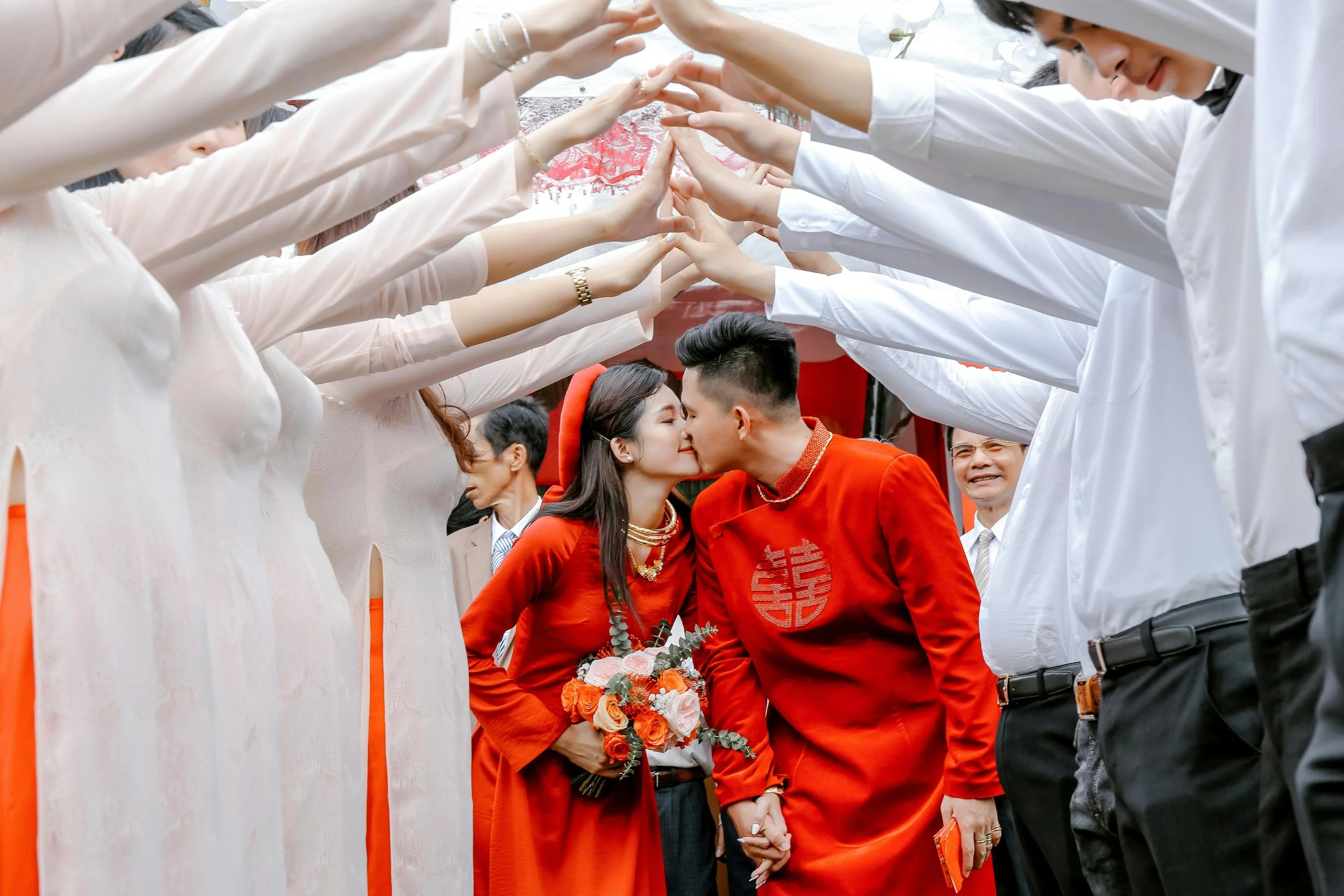 A couple dressed in traditional Vietnamese wedding attire kisses under a canopy made of raised hands during a wedding celebration.