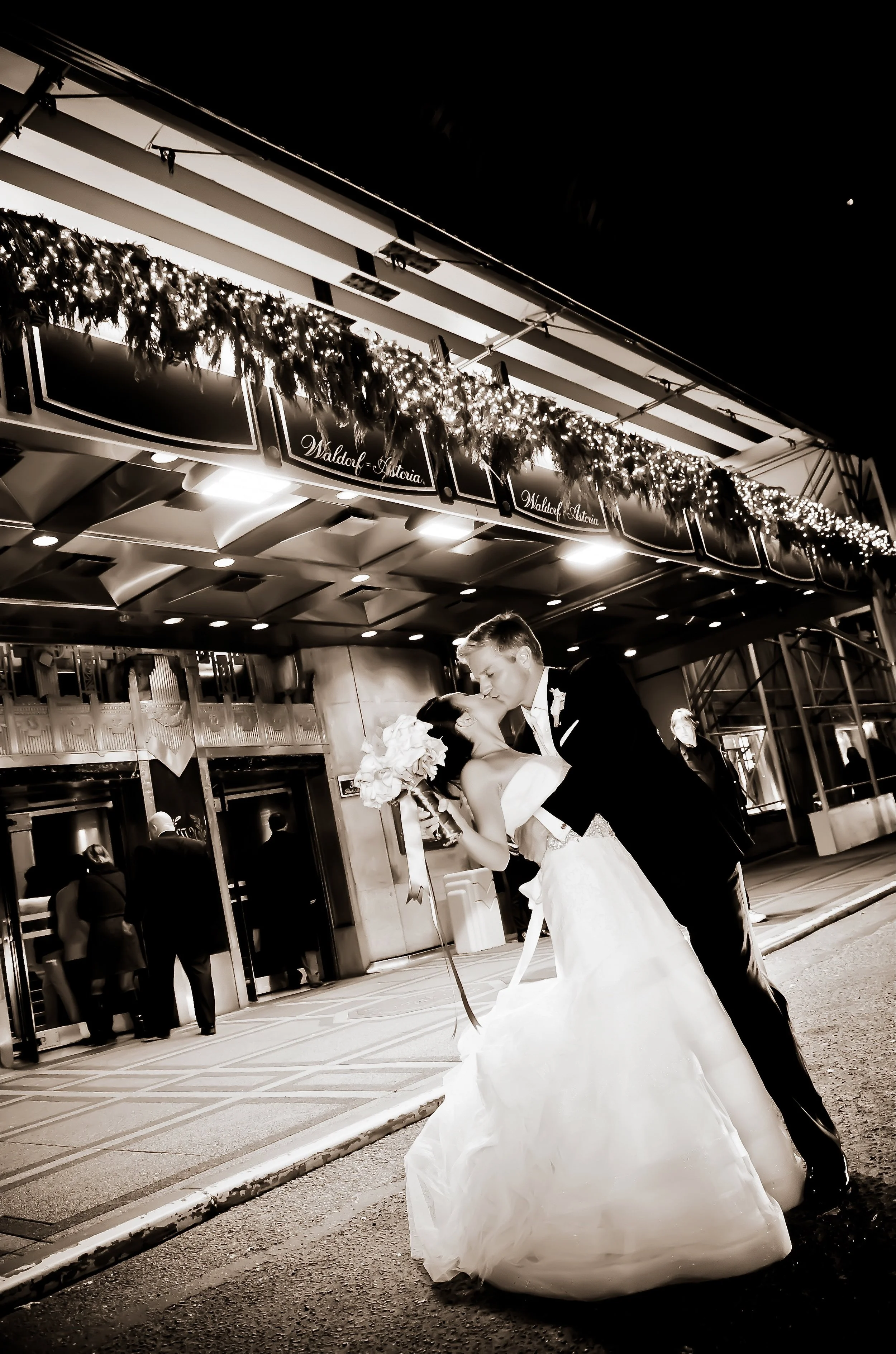 A newlywed couple sharing a kiss in front of a building decorated with holiday garland and lights at night, with a church or event venue entrance behind them.