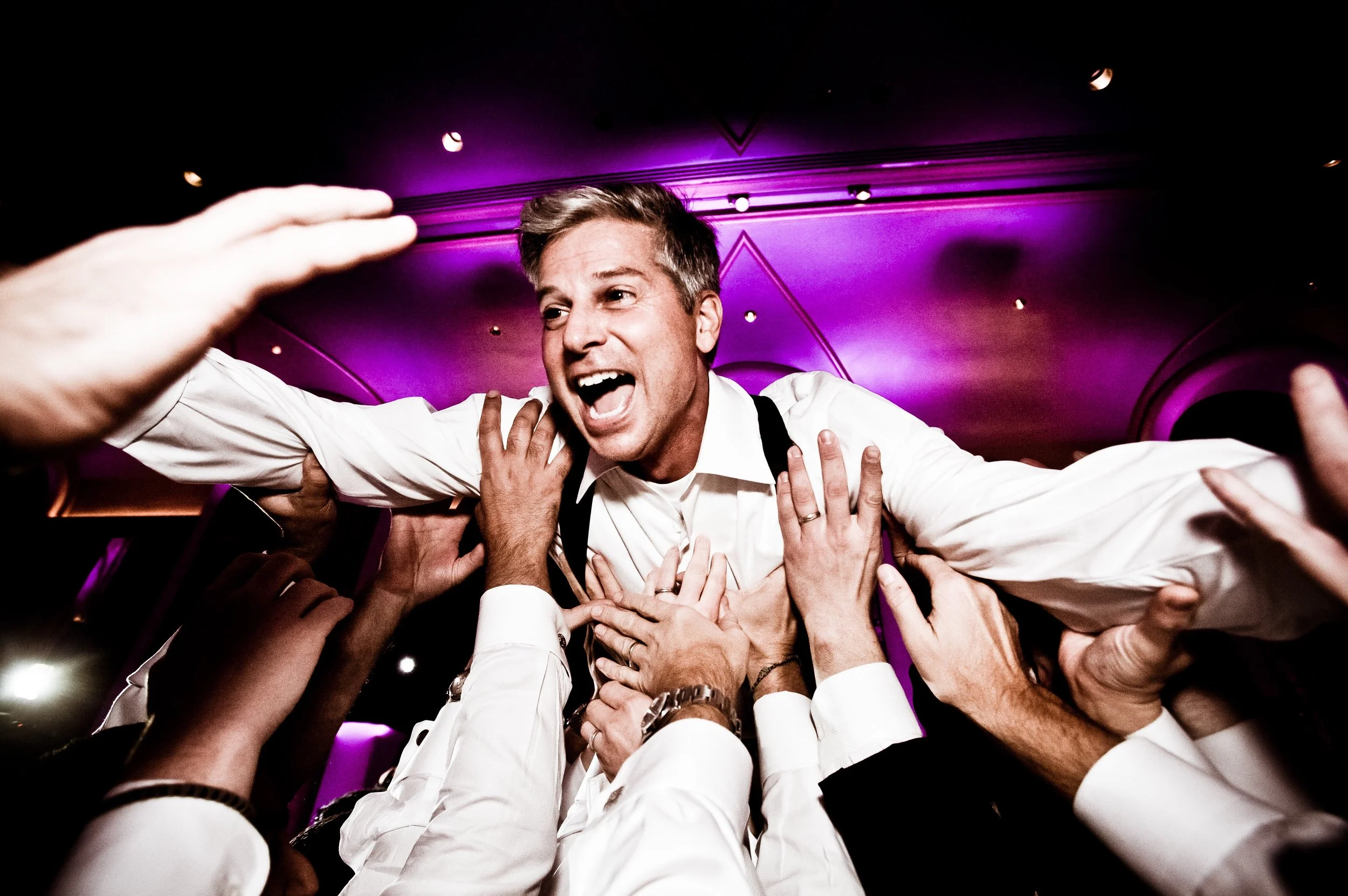 Man in a white shirt being lifted and celebrated by a group of people at a lively event with purple lighting in the background.