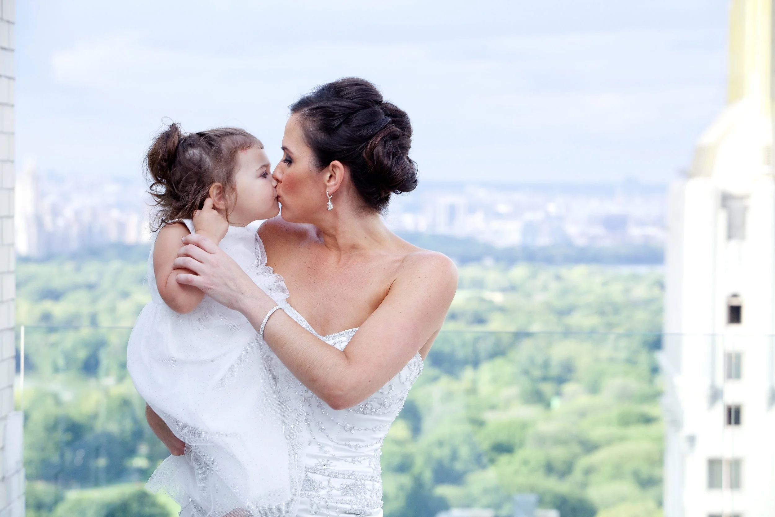 A woman in a white dress holding a young girl, both sharing a kiss on the lips with an urban cityscape background.