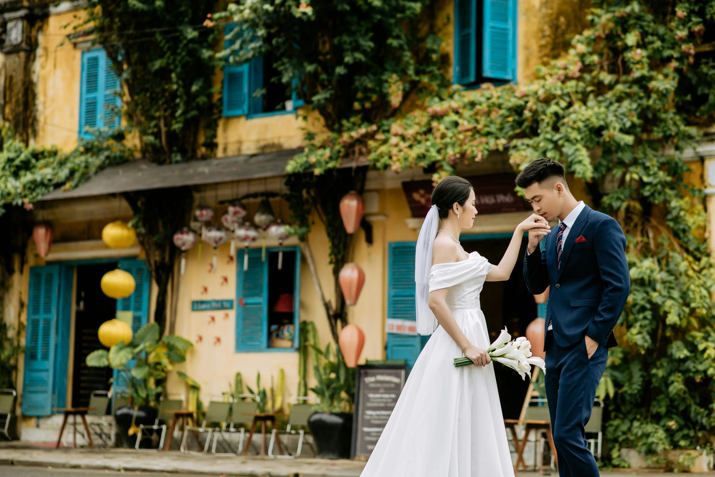 A bride and groom standing in front of a colorful building with blue shutters and hanging lanterns. The bride is holding a bouquet of white flowers, and the groom is kissing her hand.
