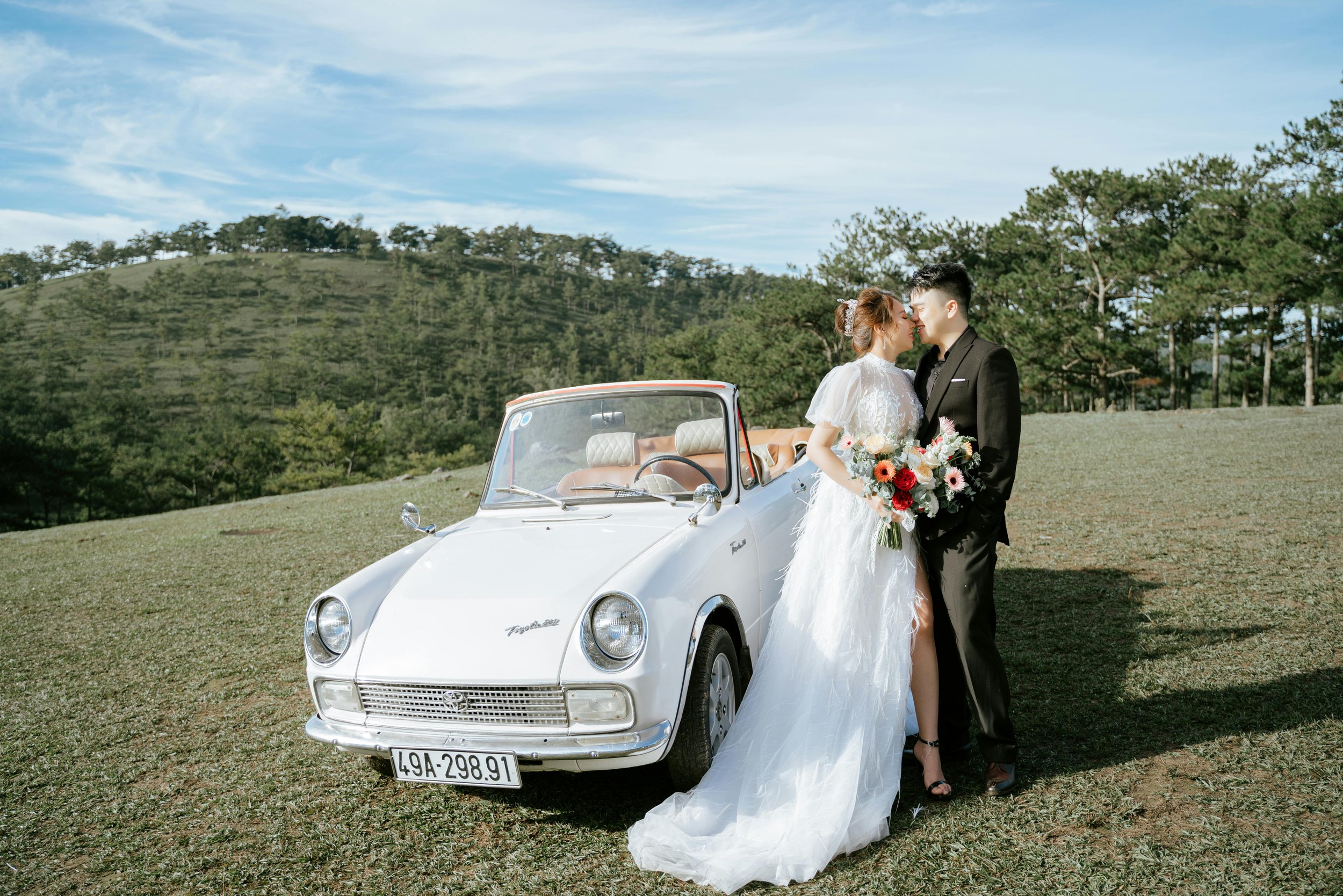 A bride and groom in wedding attire standing close together beside a vintage white convertible car on a grassy hill with trees and blue sky in the background.