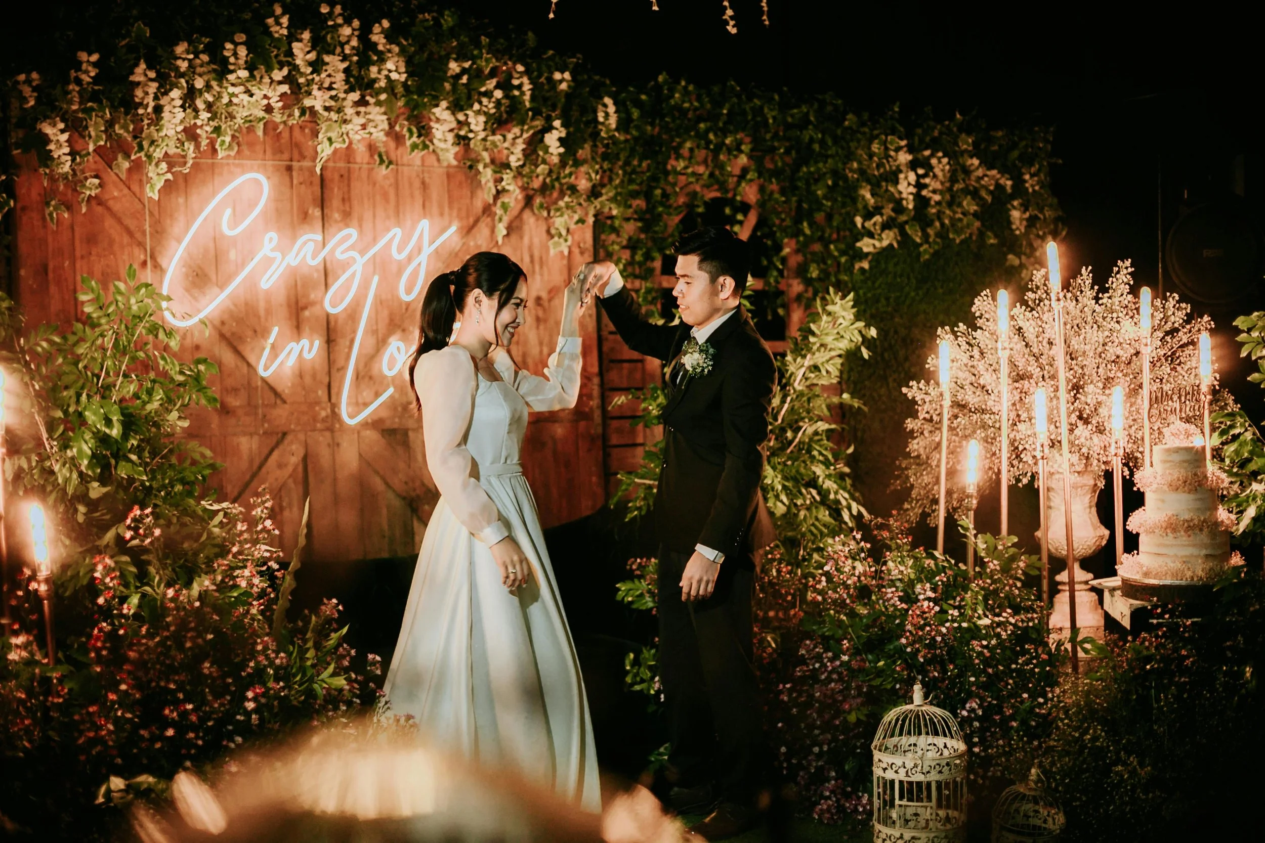 A couple in wedding attire dancing at a wedding reception with a illuminated sign that reads "Crazy in L" in the background, surrounded by flowers, foliage, candles, and decorative elements.