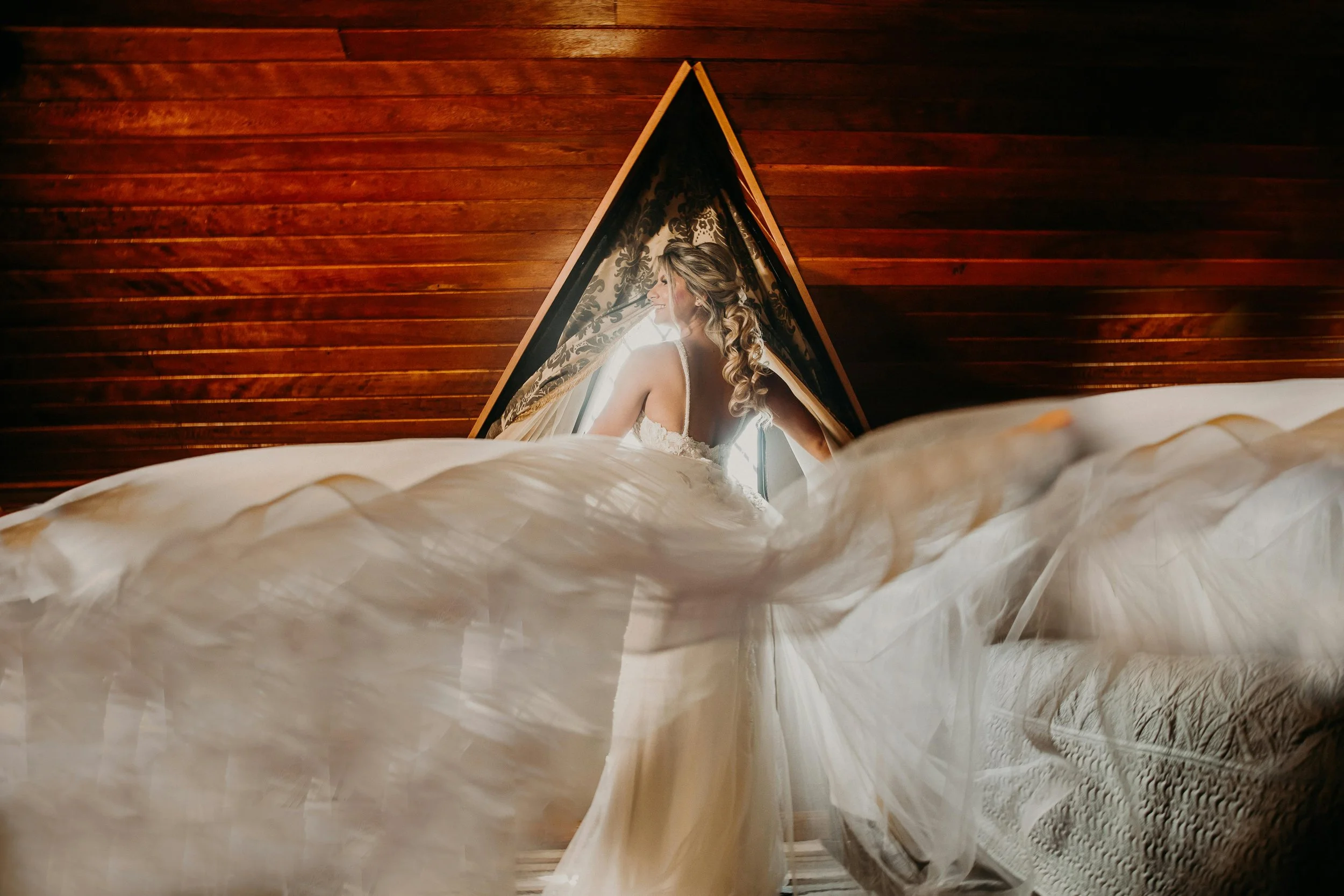 A bride in a wedding dress standing near a window with flowing curtains, with a wooden wall in the background.