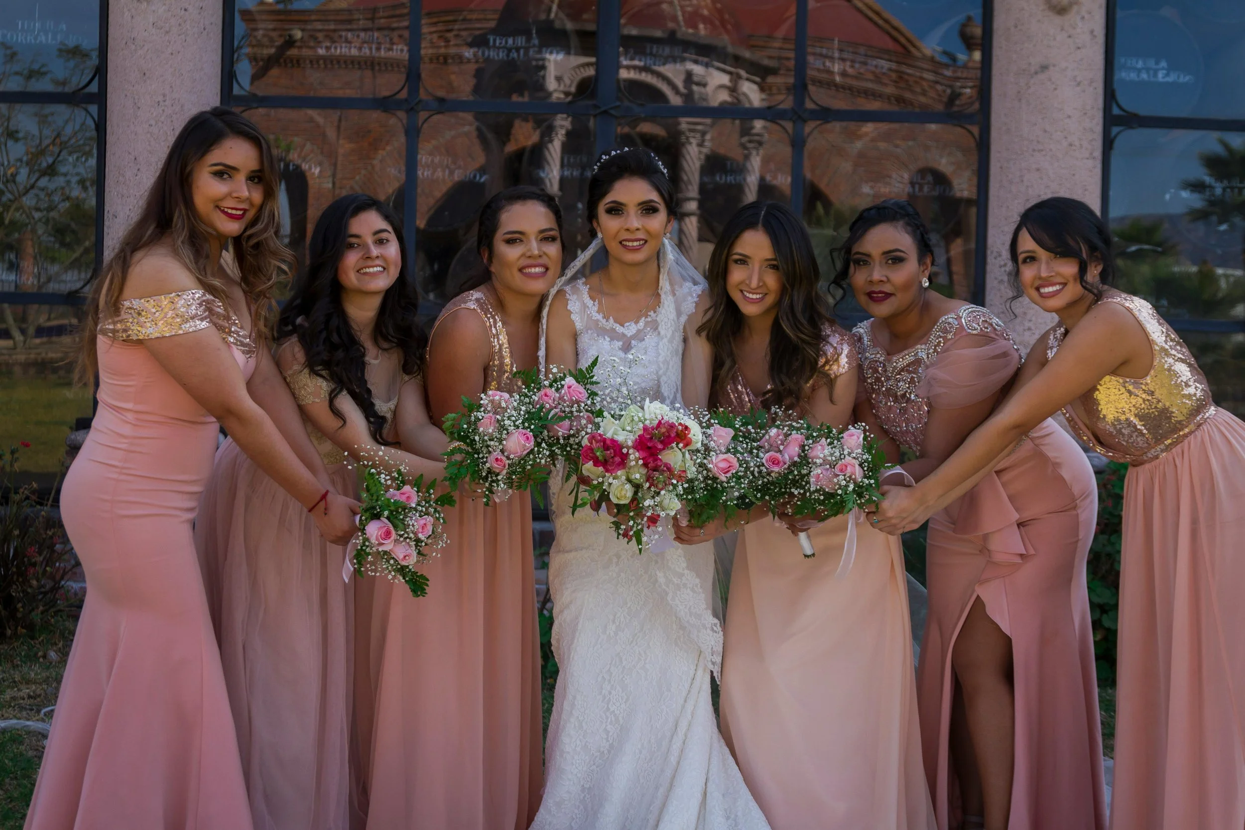 Bridal party celebrating a wedding, with the bride in a white lace wedding gown and seven bridesmaids in pink dresses holding bouquets of pink and white flowers, standing outdoors in front of a glass window reflecting an old brick building.