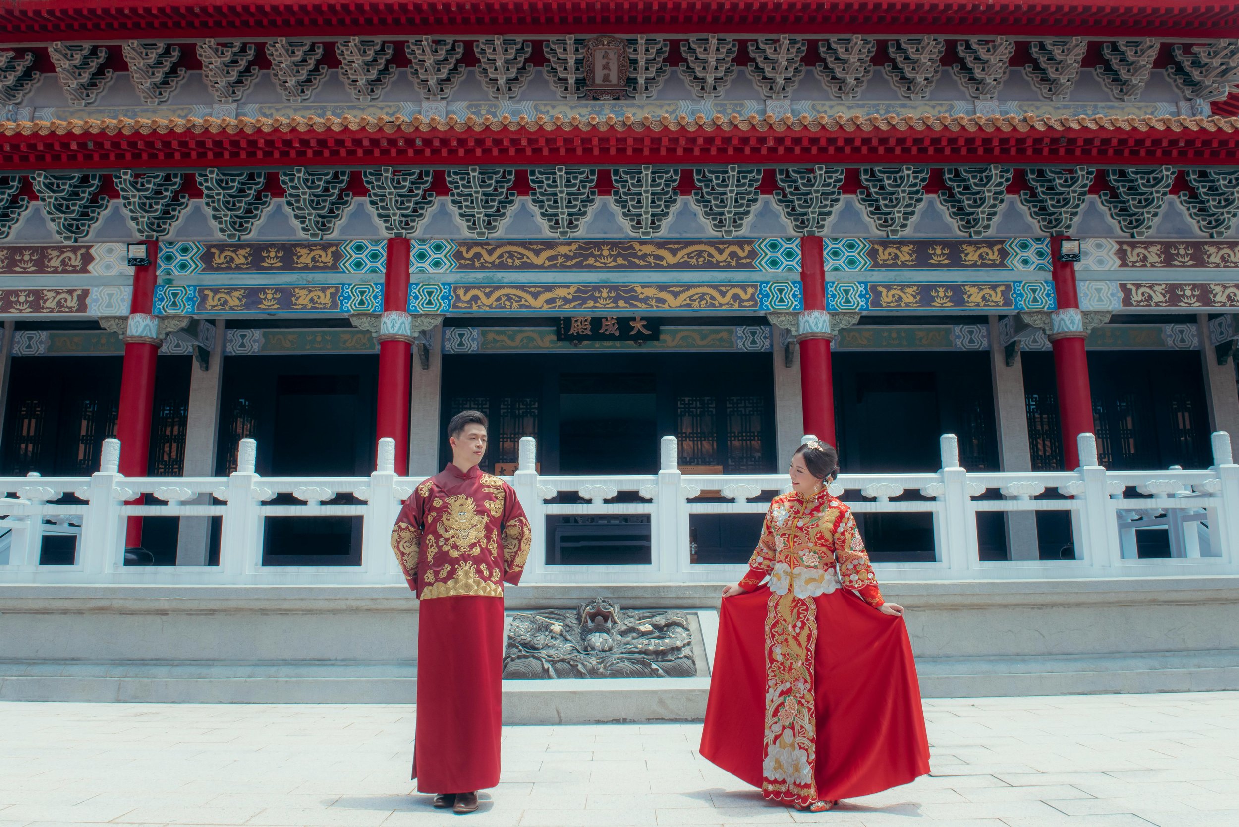 A man and woman in traditional Chinese wedding attire standing in front of an ornate historical building with red pillars, colorful decorative roof, and white fence. The woman is holding her dress, and both are looking at each other.