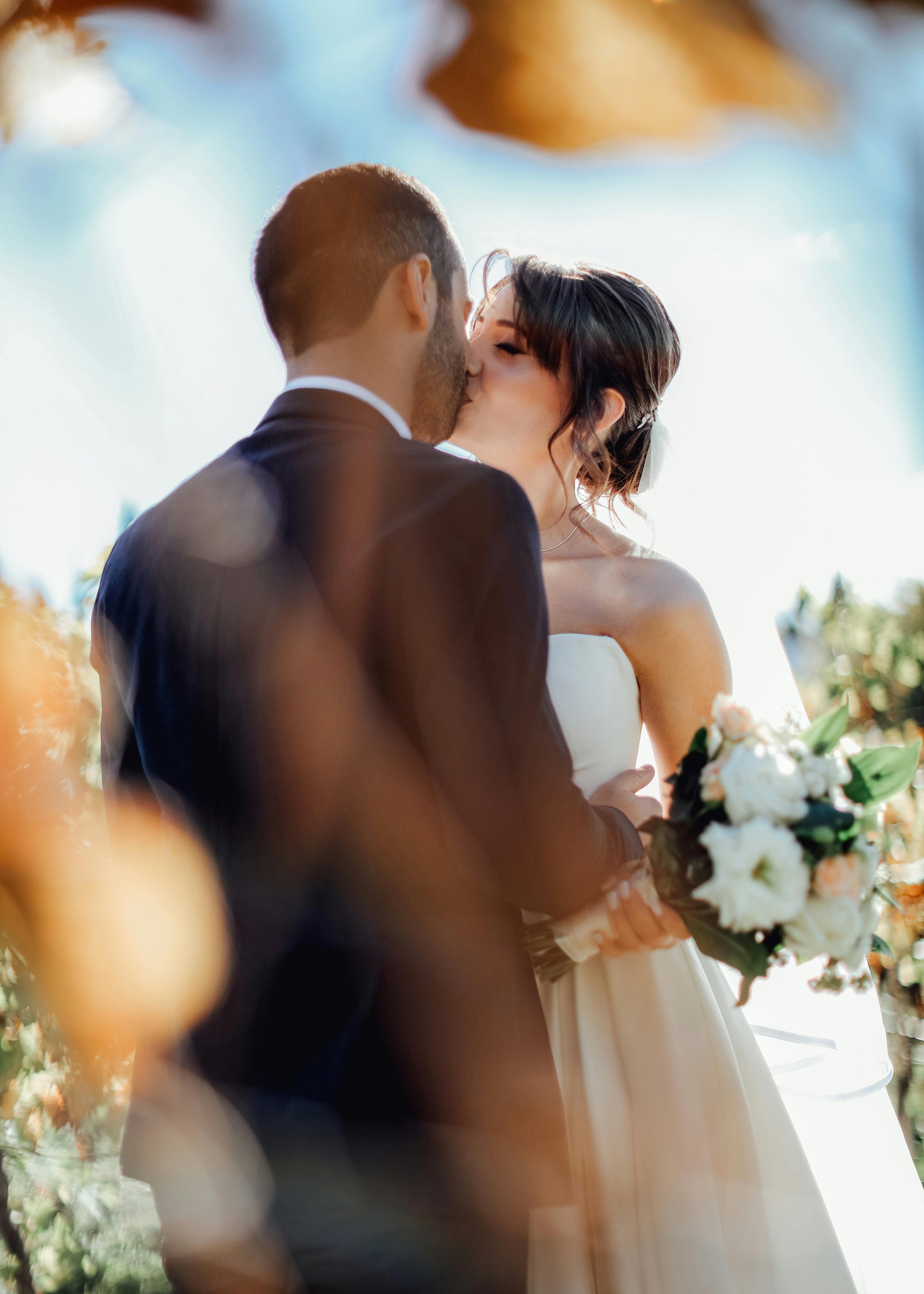 A couple kissing at their wedding, with the bride holding a bouquet of white and pink flowers and the groom in a dark suit, blurred in the foreground and background with sunlight filtering through trees.