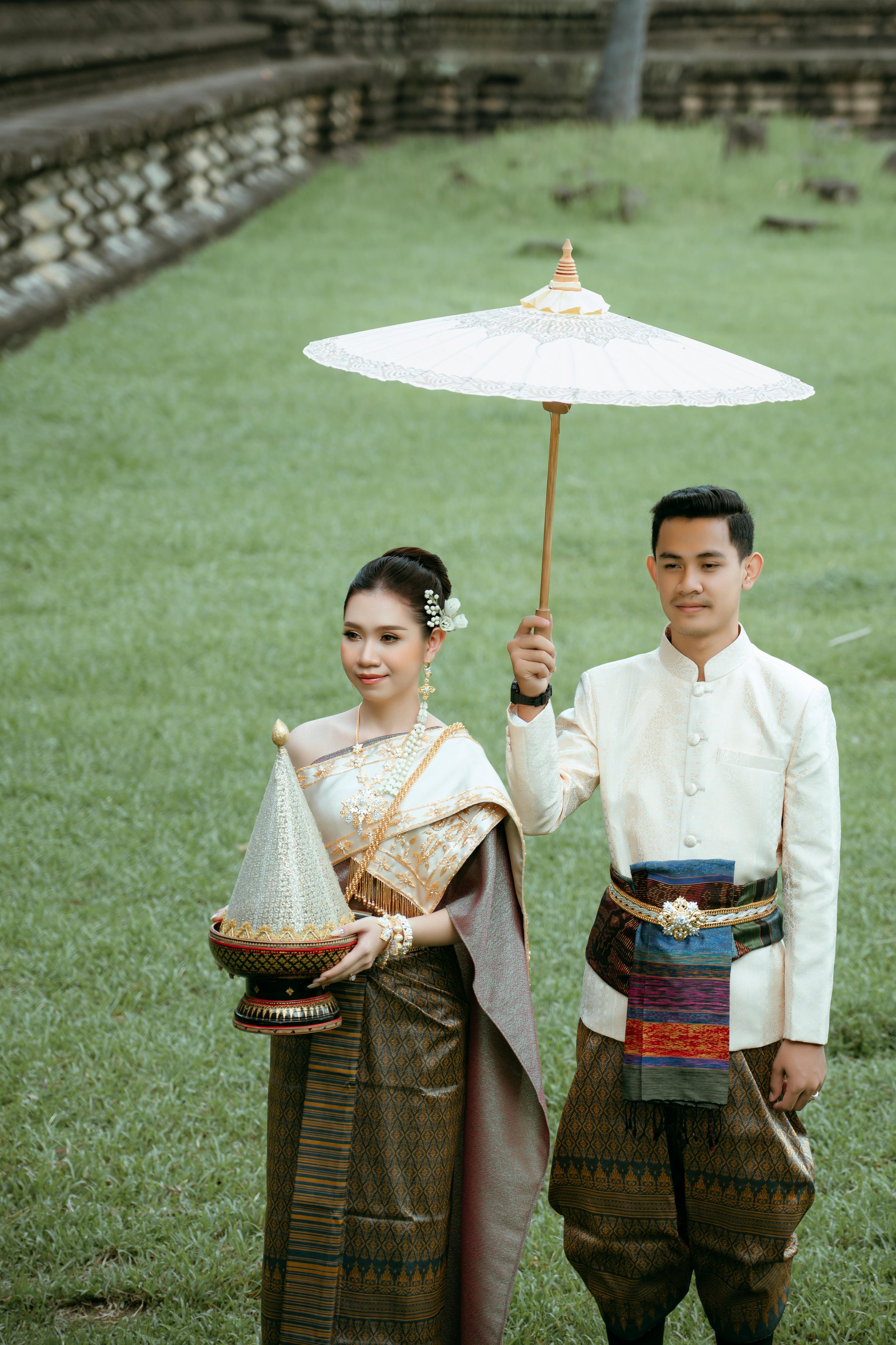 A Thai couple dressed in traditional wedding attire, with the woman holding a conical dessert and the man holding a white parasol, standing outdoors on a grassy area.