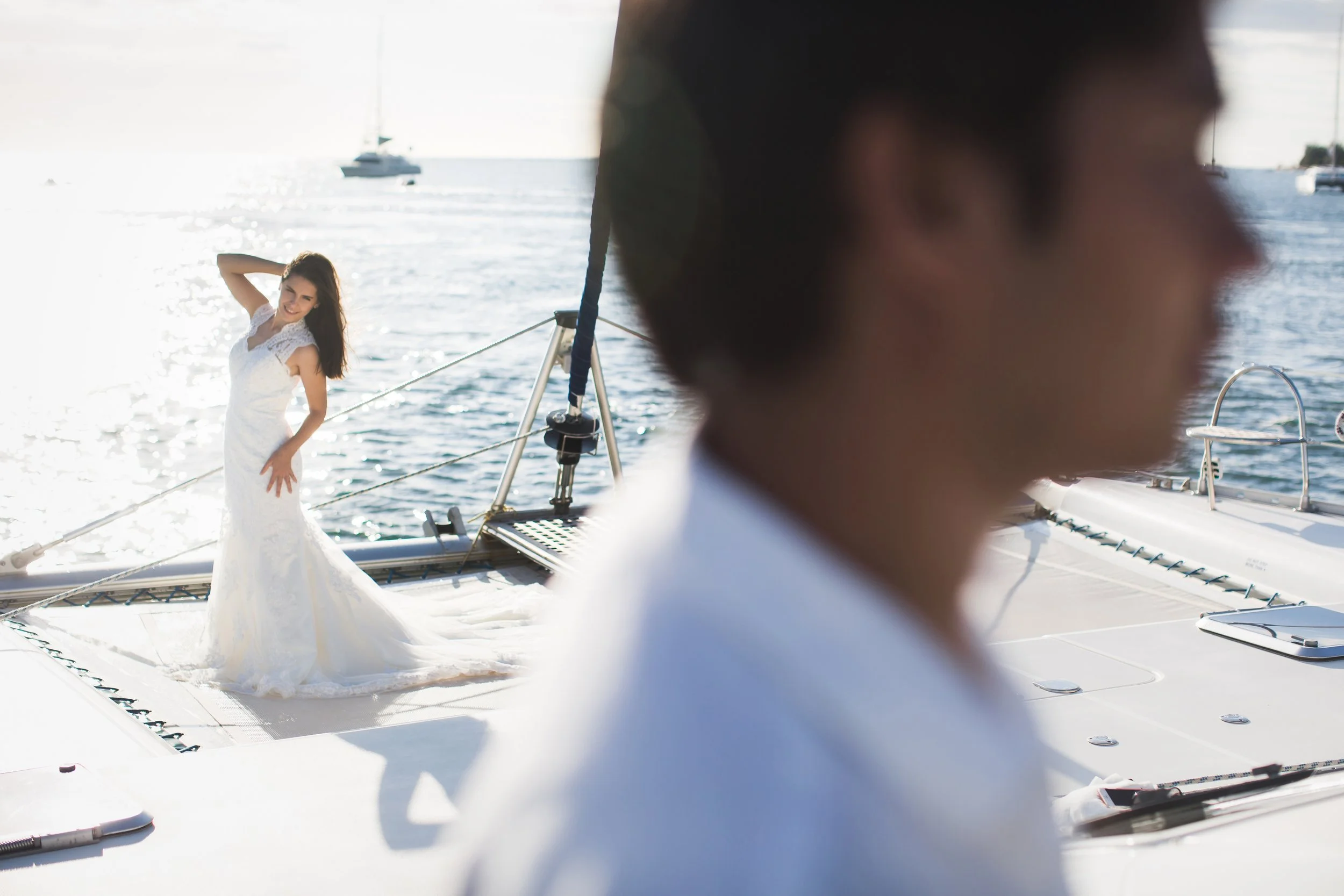 A woman in a white wedding dress standing on a boat's deck, posing with one hand on her hip, with water and sailboats in the background. A blurry man in white is in the foreground.