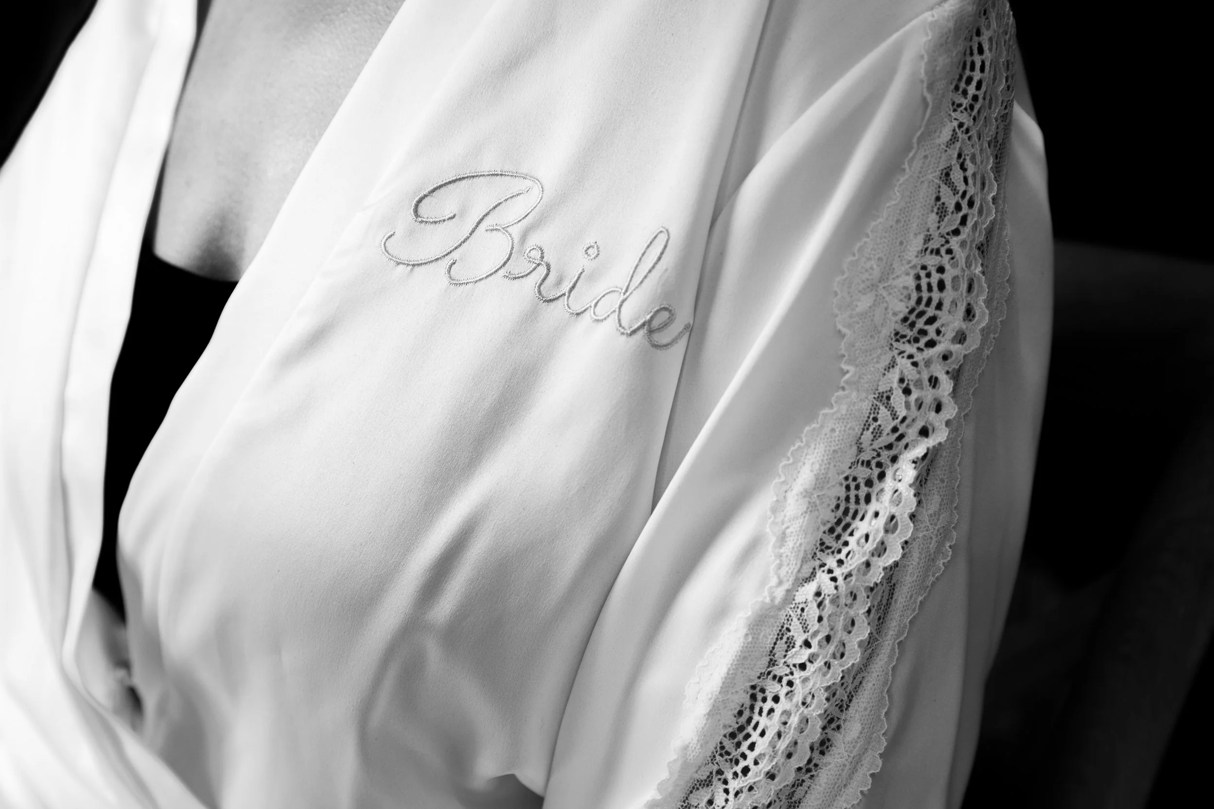 Close-up of a bride's robe featuring lace details on the sleeve and embroidered text 'Bride' on the front in black and white.
