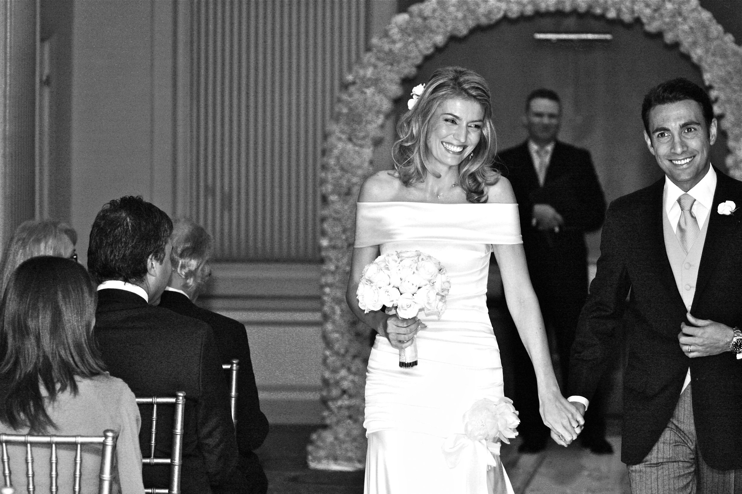 A bride and groom smiling and holding hands during their wedding ceremony, with guests seated and a floral arch in the background.