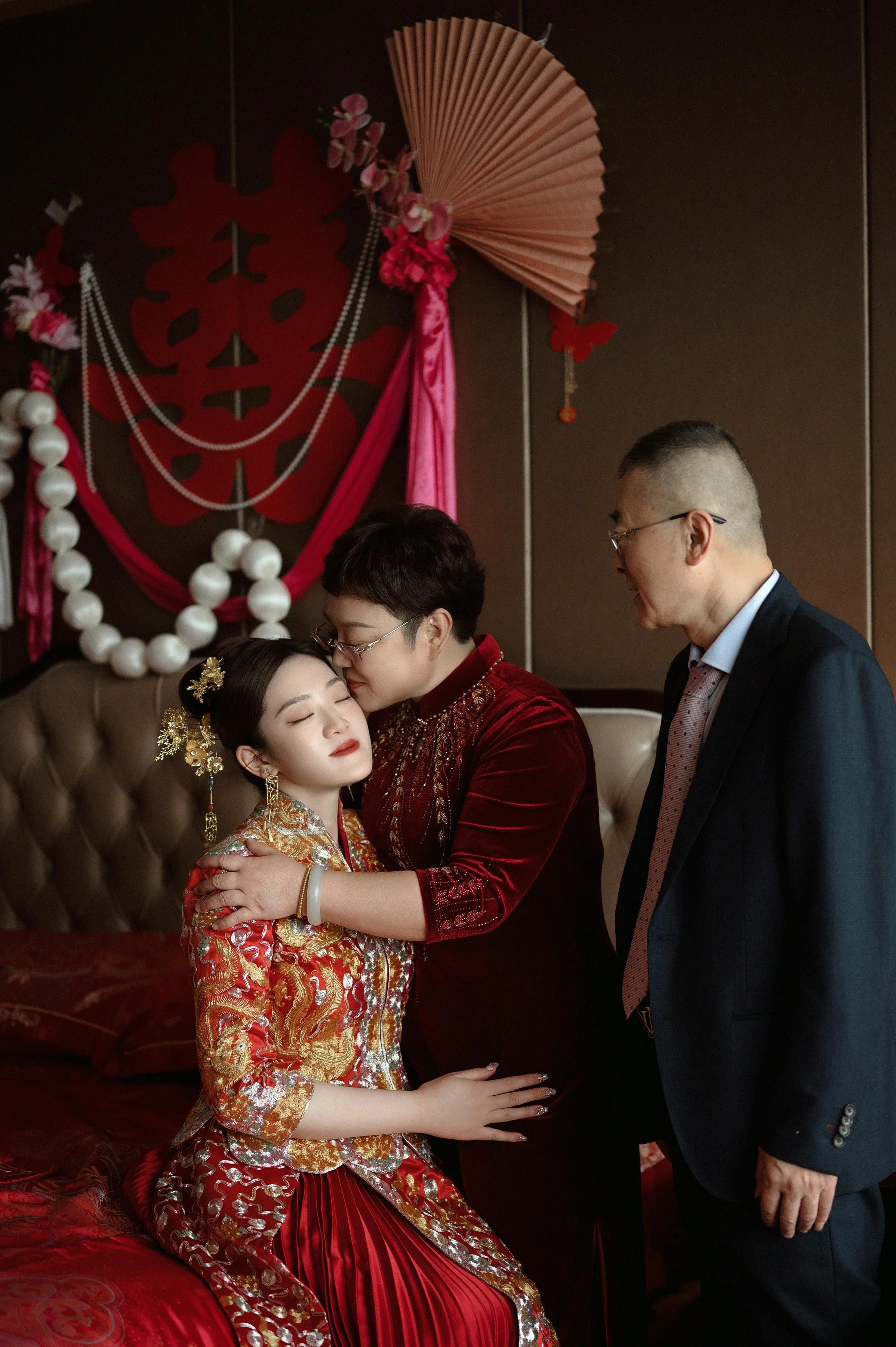A woman dressed in traditional Chinese wedding attire is being embraced and kissed on the forehead by an older woman, possibly her mother, in a decorated room with Chinese-themed decorations and a man in a suit watching them.