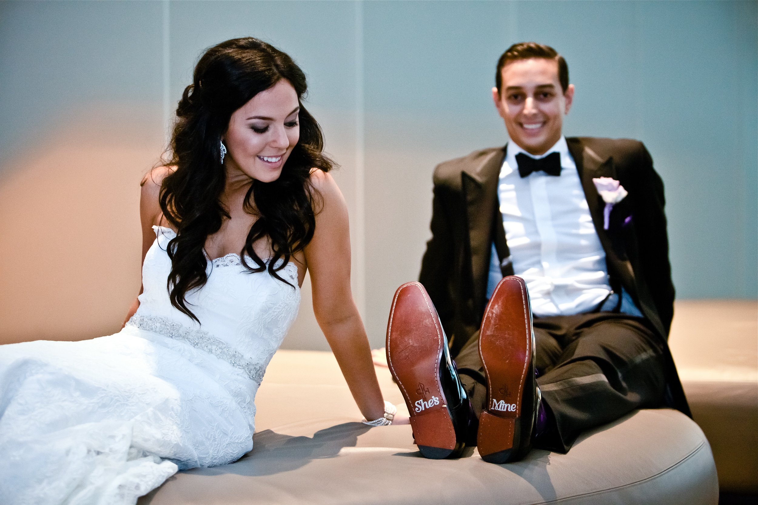 A bride and groom sitting on a bed, smiling and looking at each other, with their shoes in front of them. The bride is wearing a white wedding dress, and the groom is in a tuxedo with a bowtie.