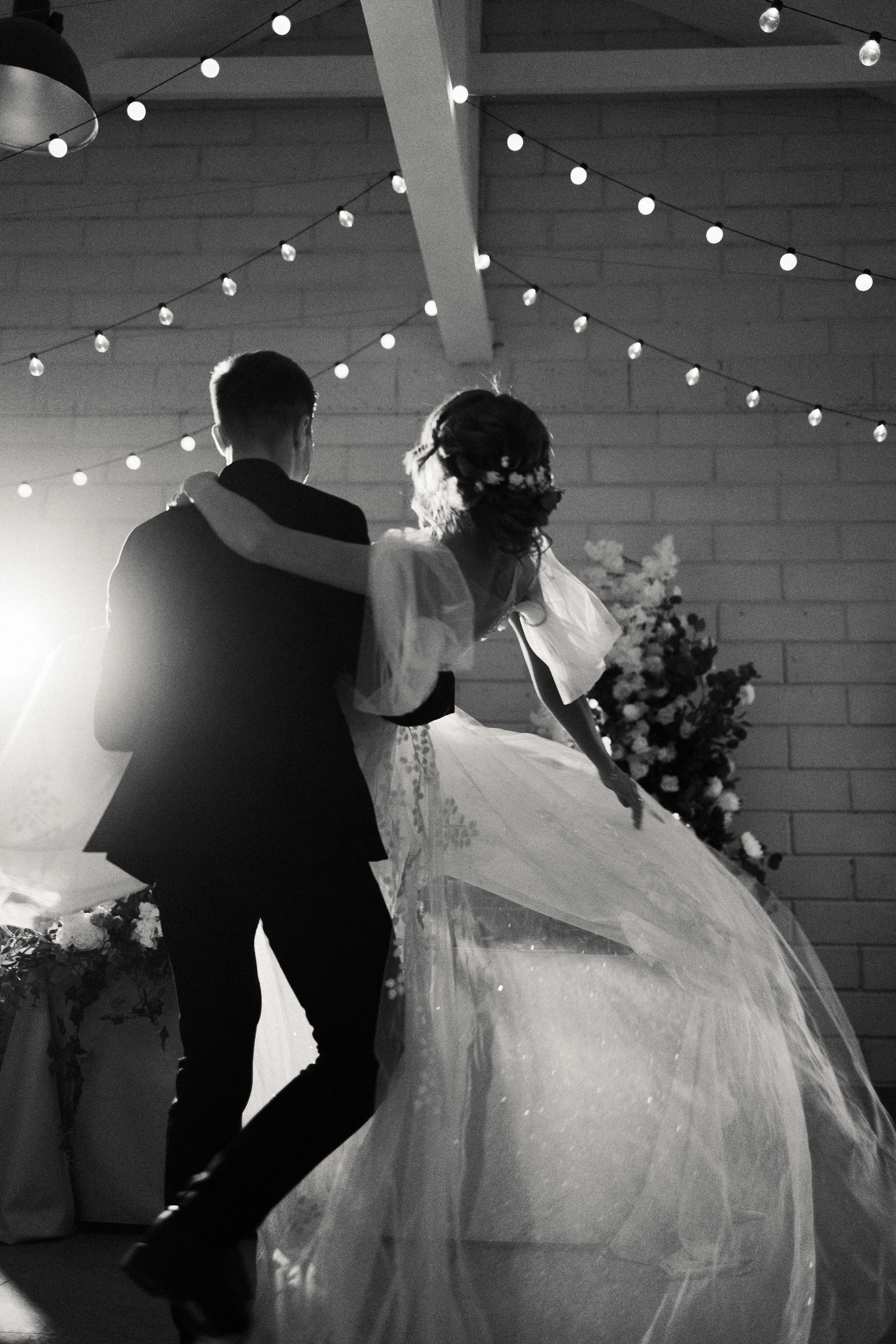 A black and white photo of a bride and groom dancing at their wedding reception, with string lights hanging from the ceiling.
