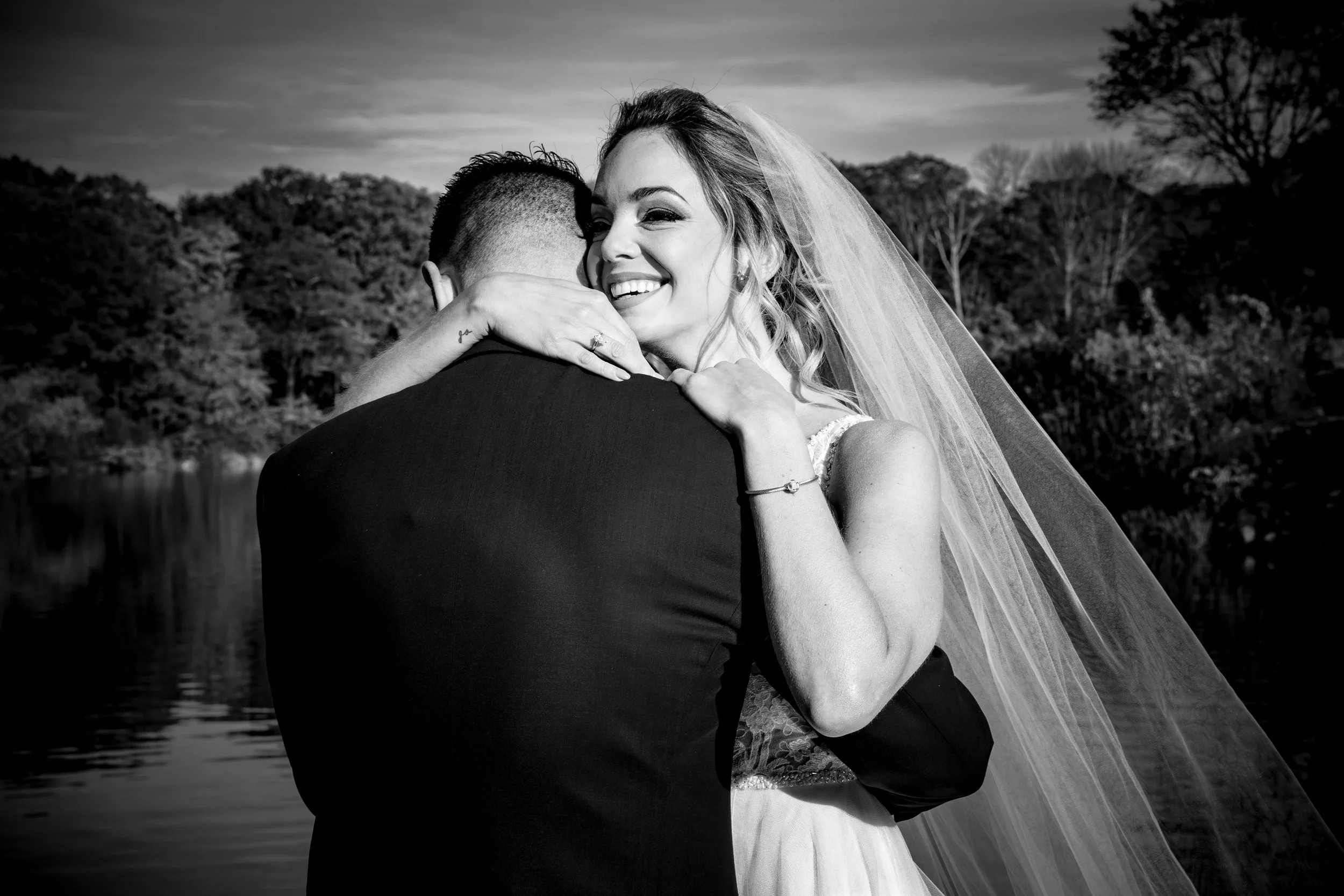 A black-and-white photo of a bride and groom embracing outdoors near a body of water with trees in the background. The bride is smiling, wearing a wedding dress and veil. The groom, in a dark suit, is facing away from the camera.
