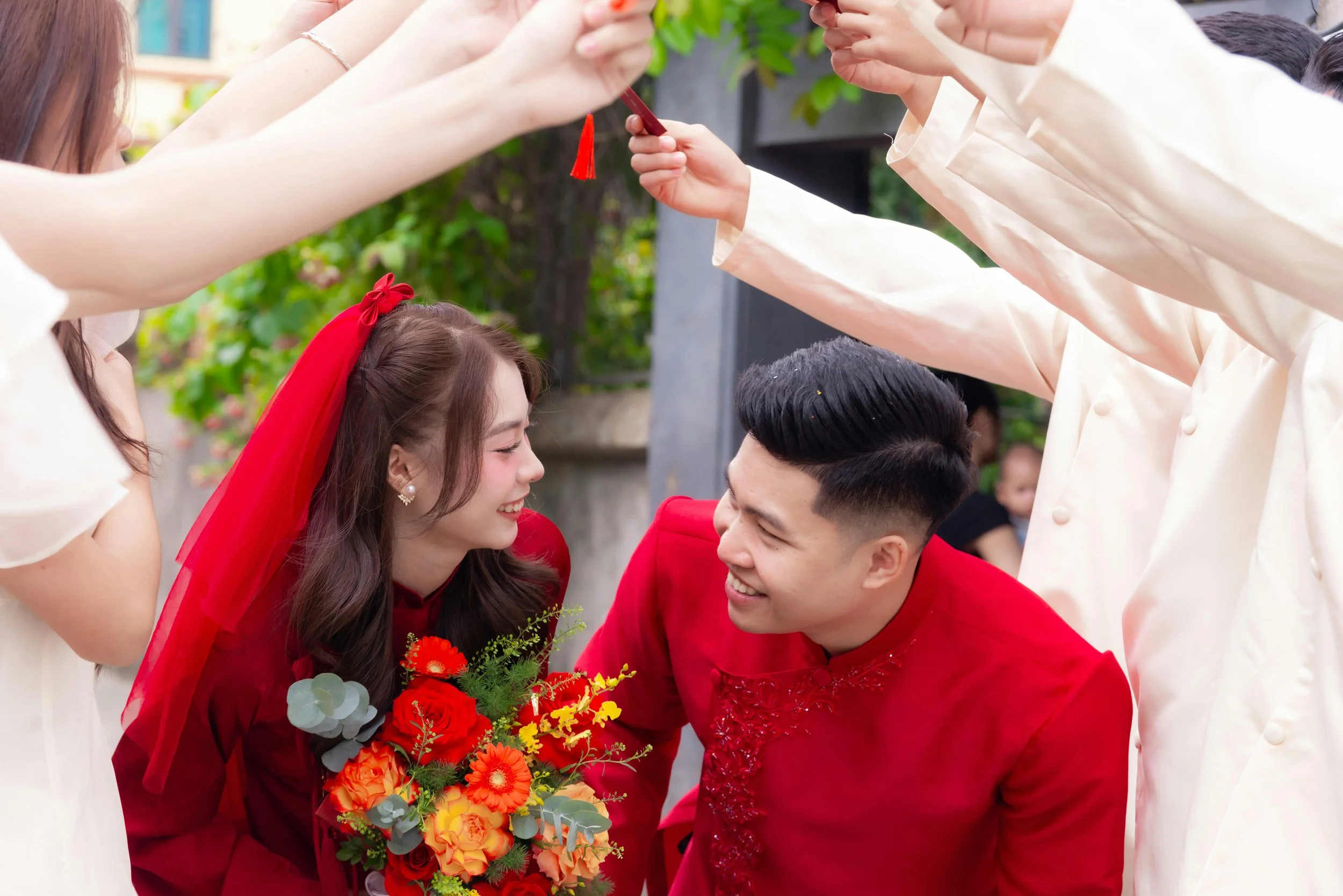 A couple dressed in traditional red attire exchange vows during a wedding ceremony with family members holding a red thread over their heads.
