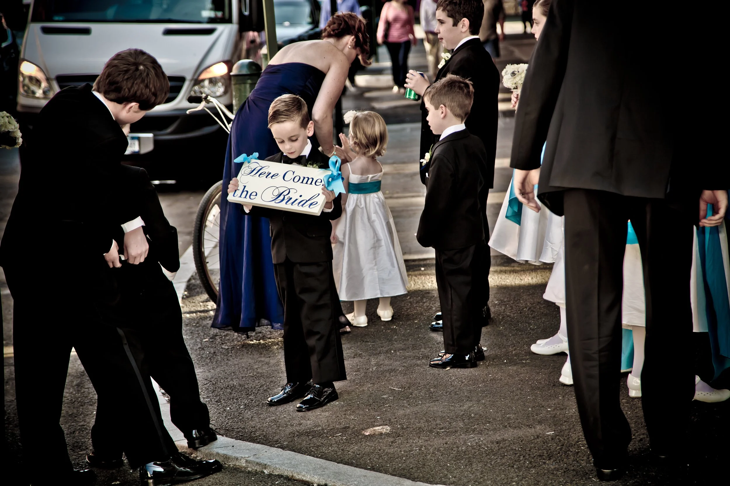 Group of children in formal attire, some holding flowers, standing on a street in a wedding procession. One child holds a sign saying 'Here Comes the Bride,' with cars parked in the background.