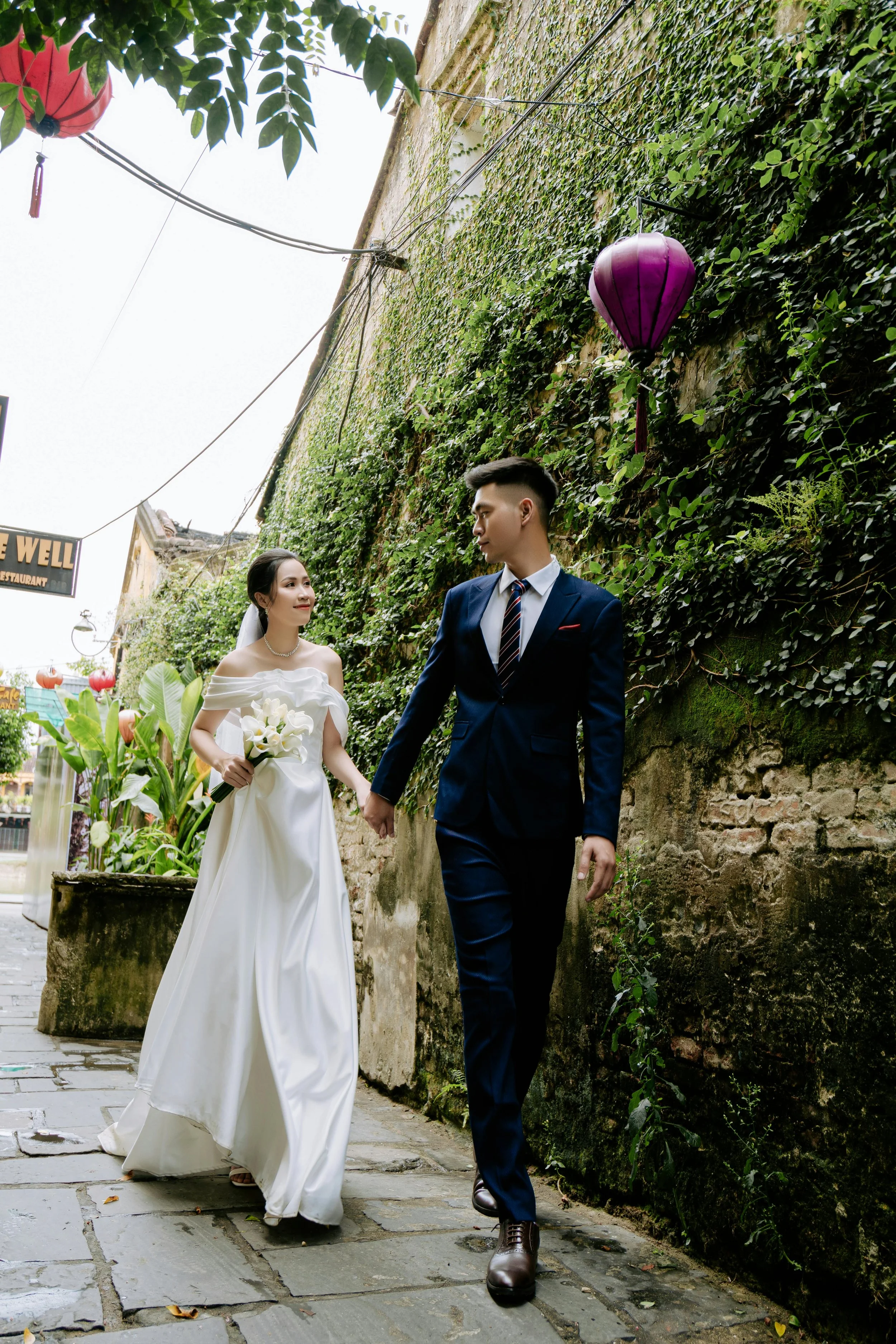 A bride and groom holding hands and walking on a cobblestone street, surrounded by greenery and colorful lanterns.