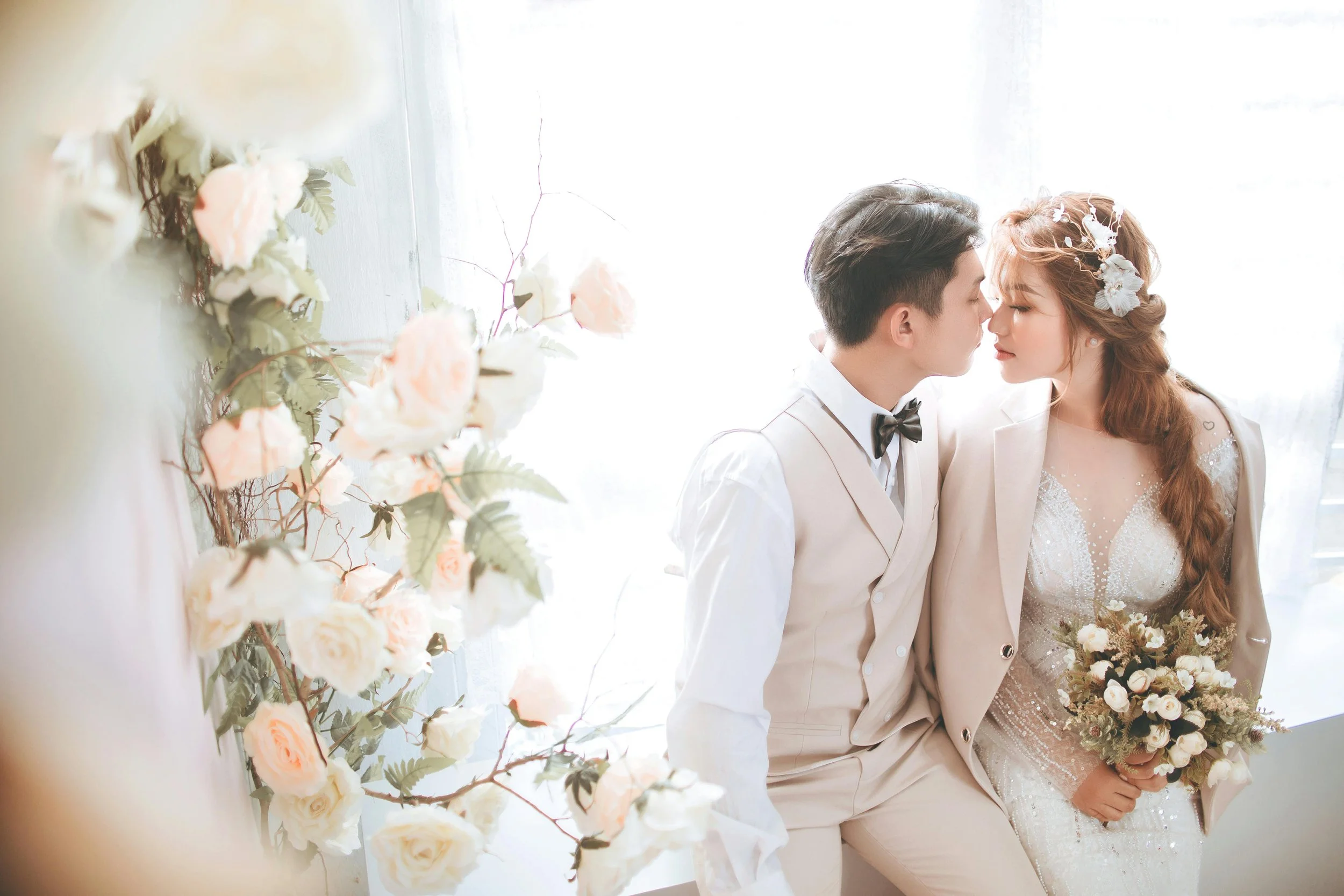 A newlywed couple leaning in for a kiss in a softly lit room, with a bouquet of white flowers and greenery, and decorative flowers in their hair, wearing elegant wedding attire.