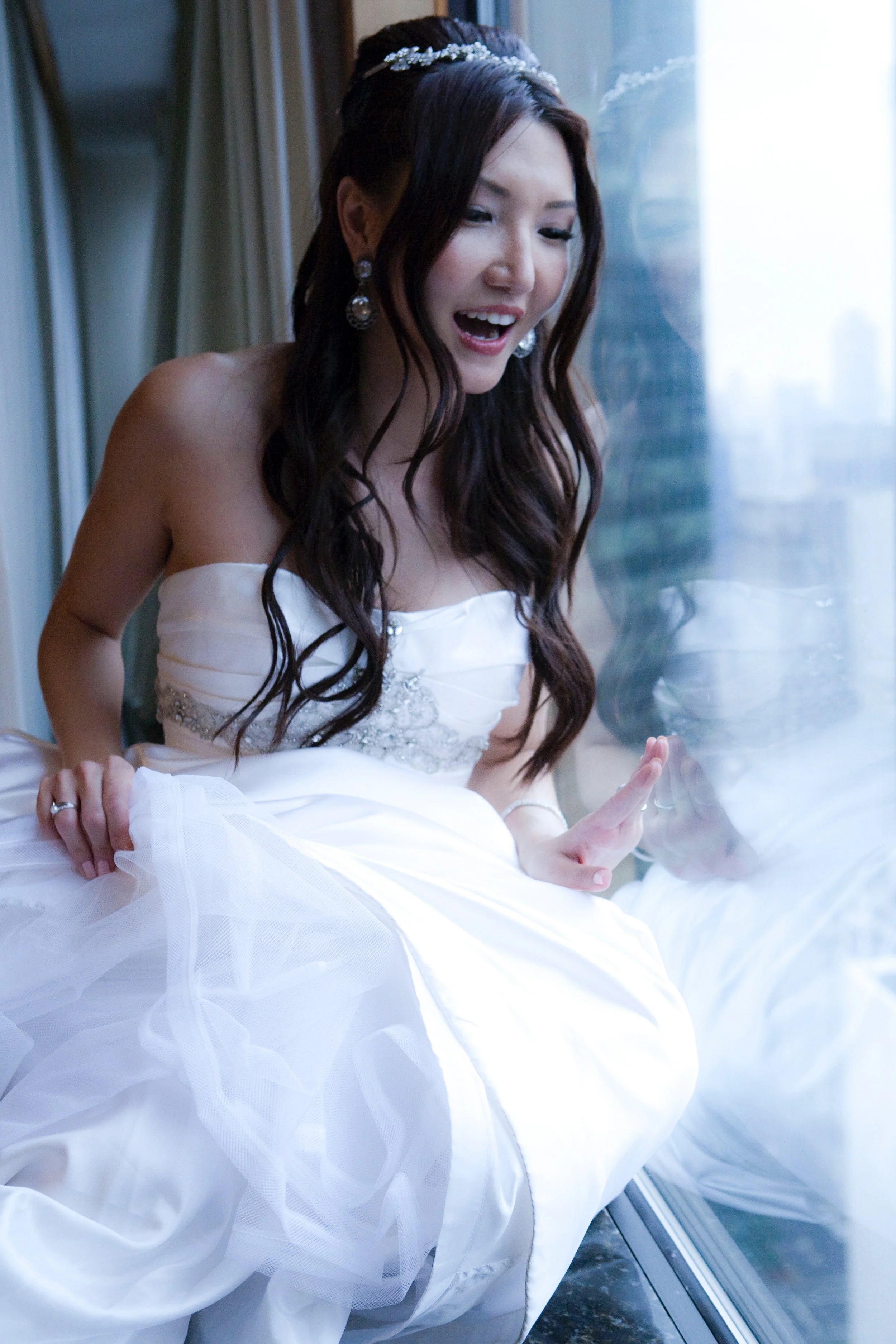 A woman in a white wedding dress with dark brown hair looking at her reflection in a window.