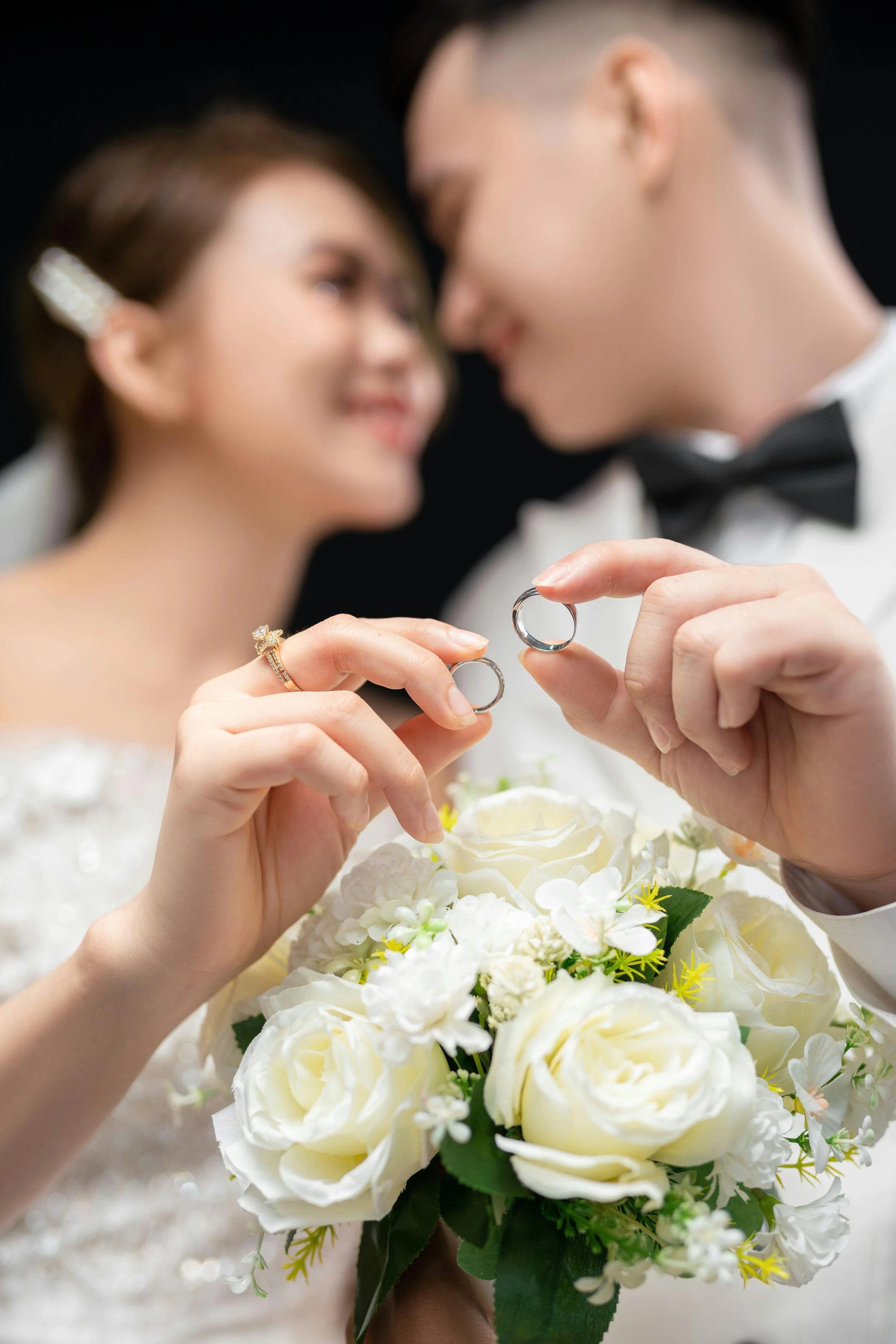 A bride and groom hold wedding rings in front of a bouquet of white roses and flowers, with their faces blurred in the background.