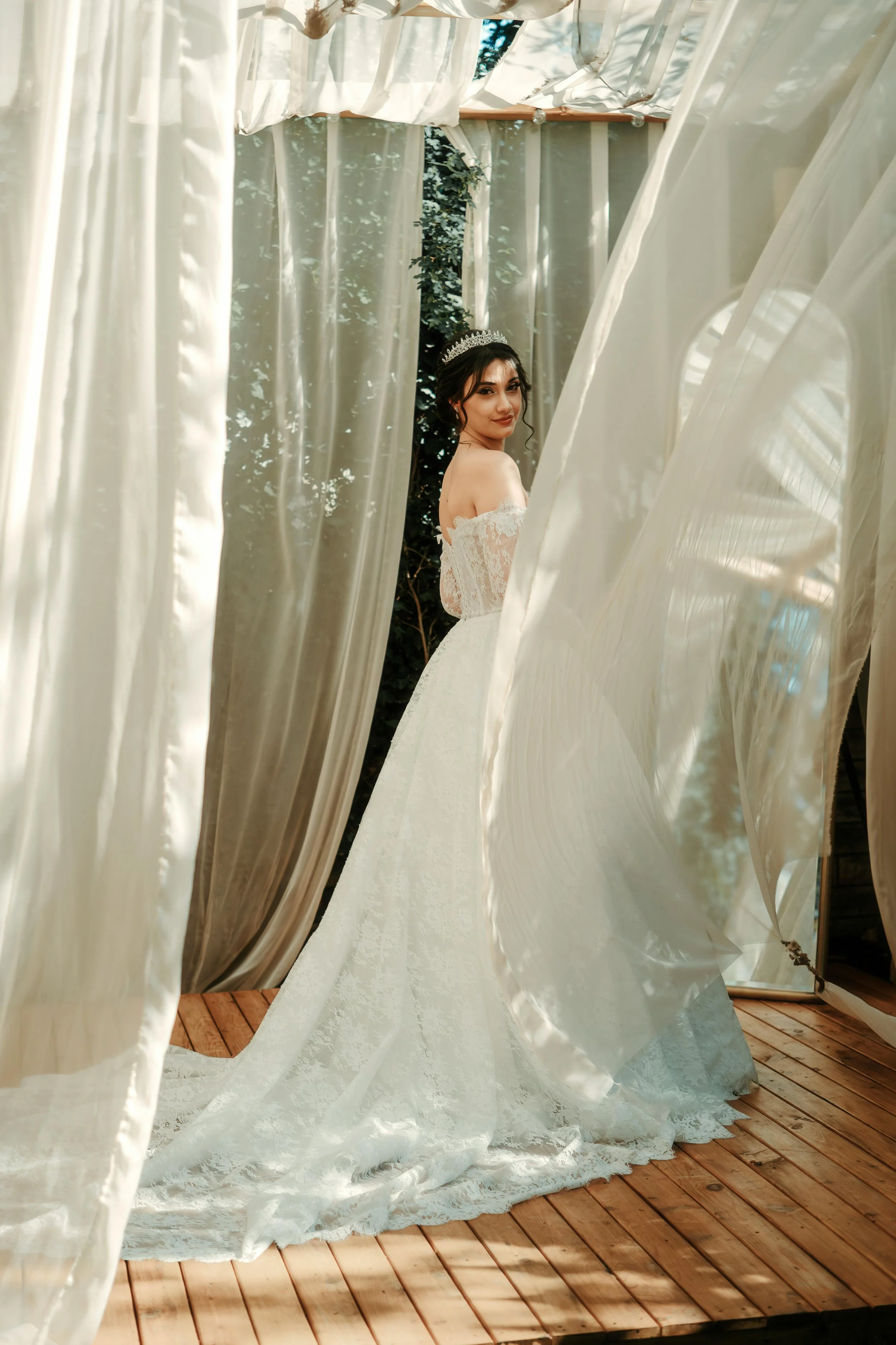 A bride in a white wedding dress with lace details, standing on a wooden floor inside a tent with cream-colored curtains, wearing a tiara and smiling while looking over her shoulder.
