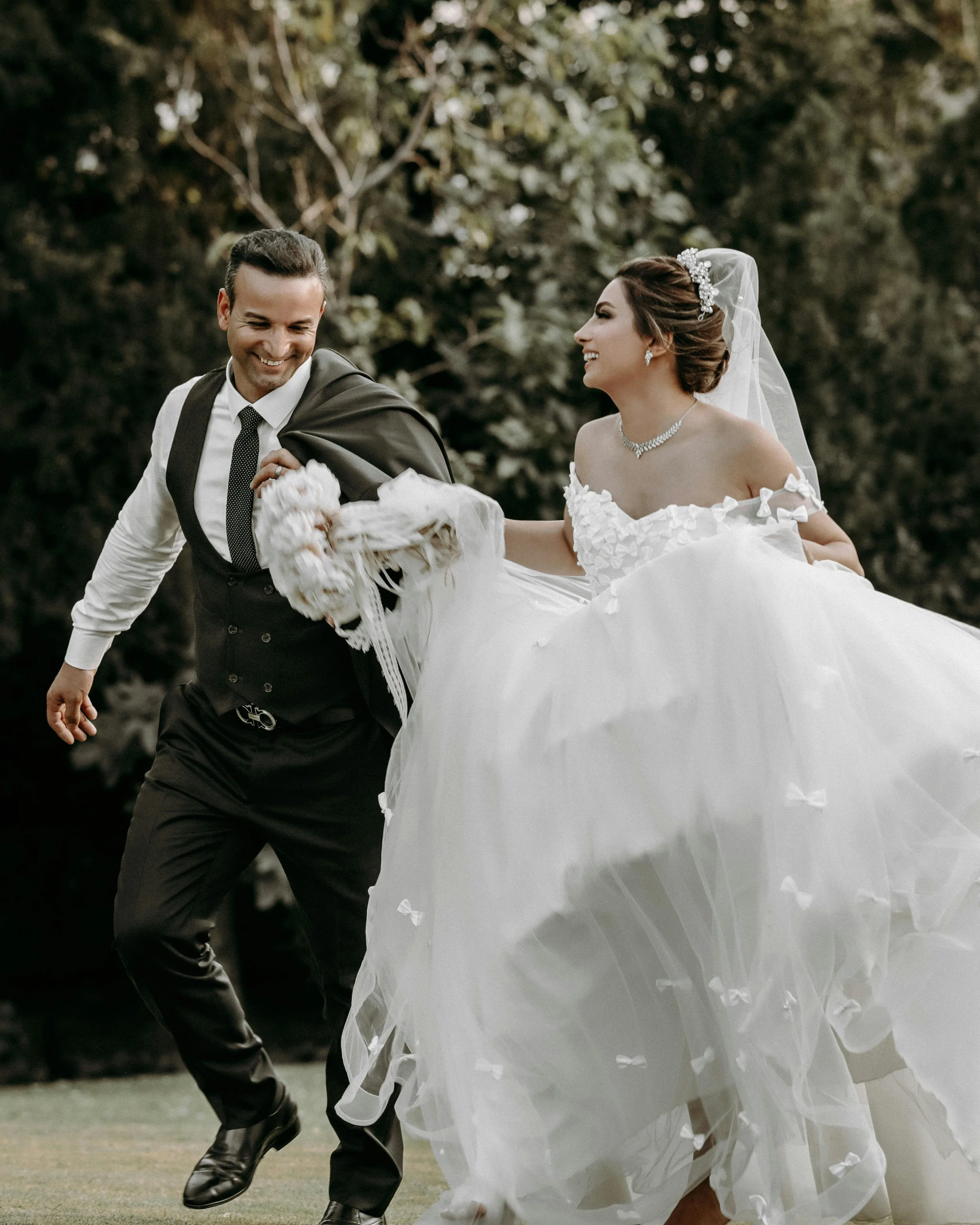 A newlywed couple walking outdoors, with the groom in a suit and the bride in a white wedding gown with a veil, both smiling and happy.