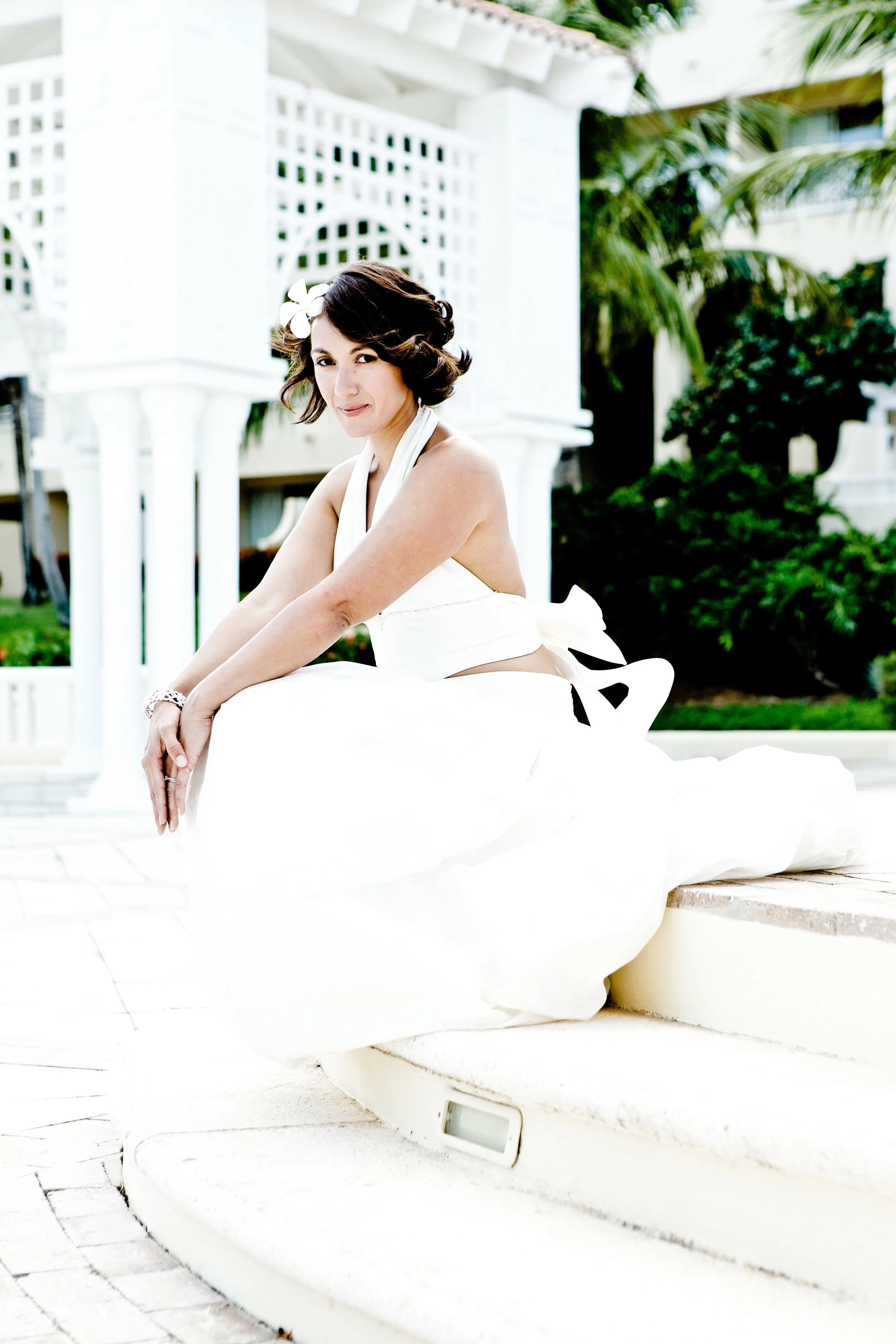 A woman in a white dress with a flower in her hair, sitting outdoors on steps with white architecture and greenery in the background.
