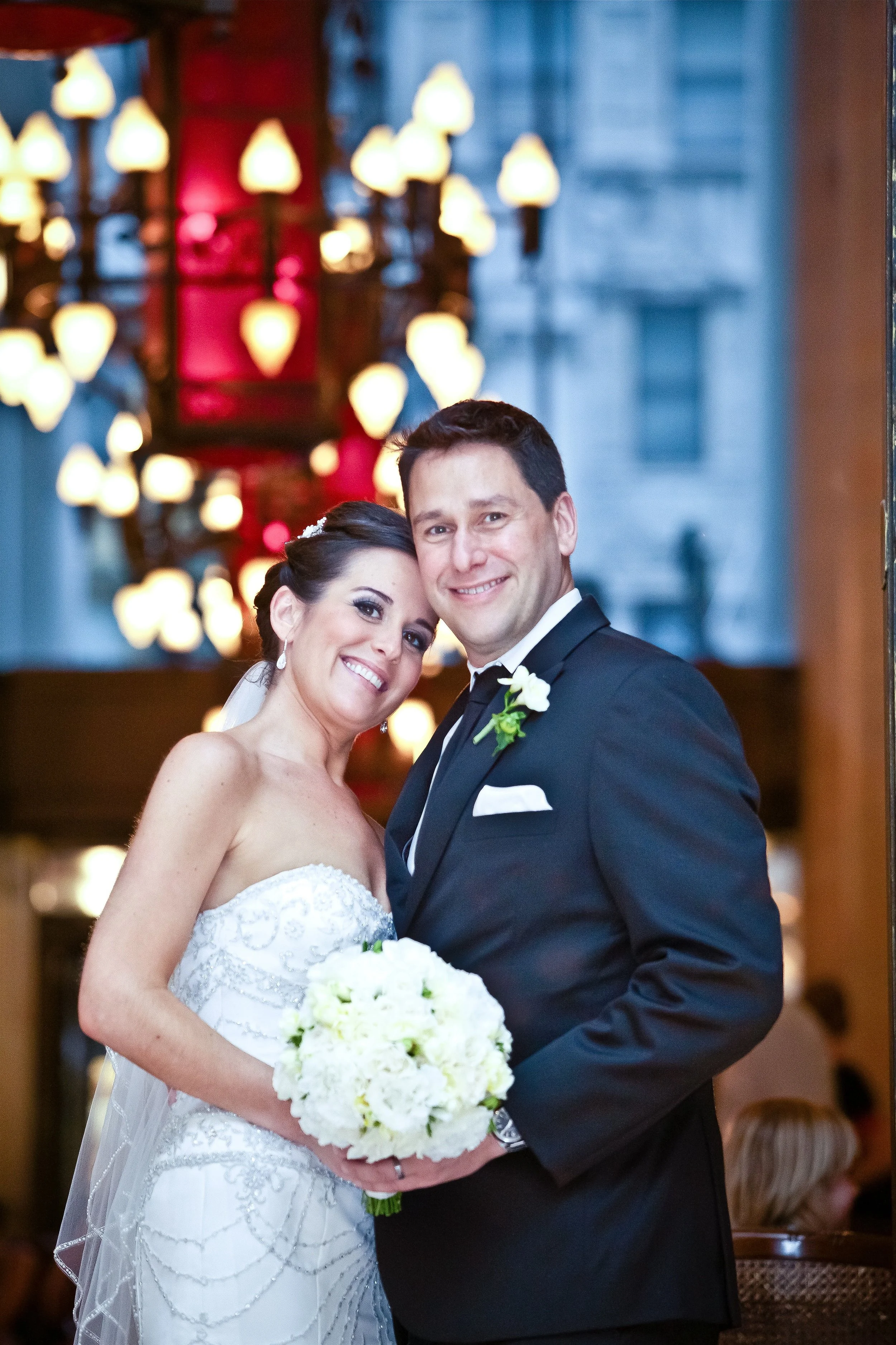 A bride and groom smiling and posing for a picture indoors with a chandelier in the background.