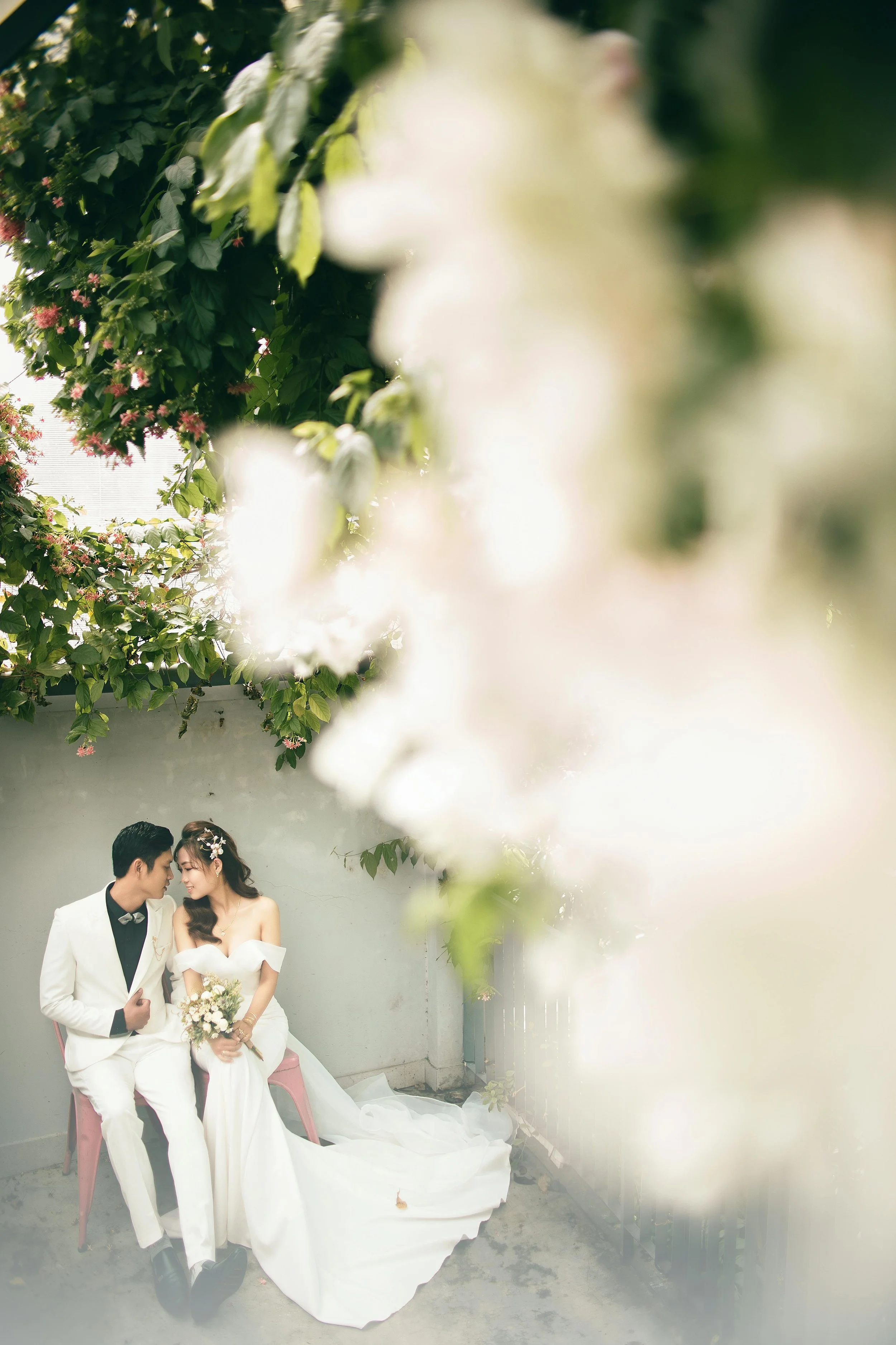A bride and groom sitting close together on pink chairs indoors, with white flowers in her hair and holding a bouquet, surrounded by greenery and white blossoms.