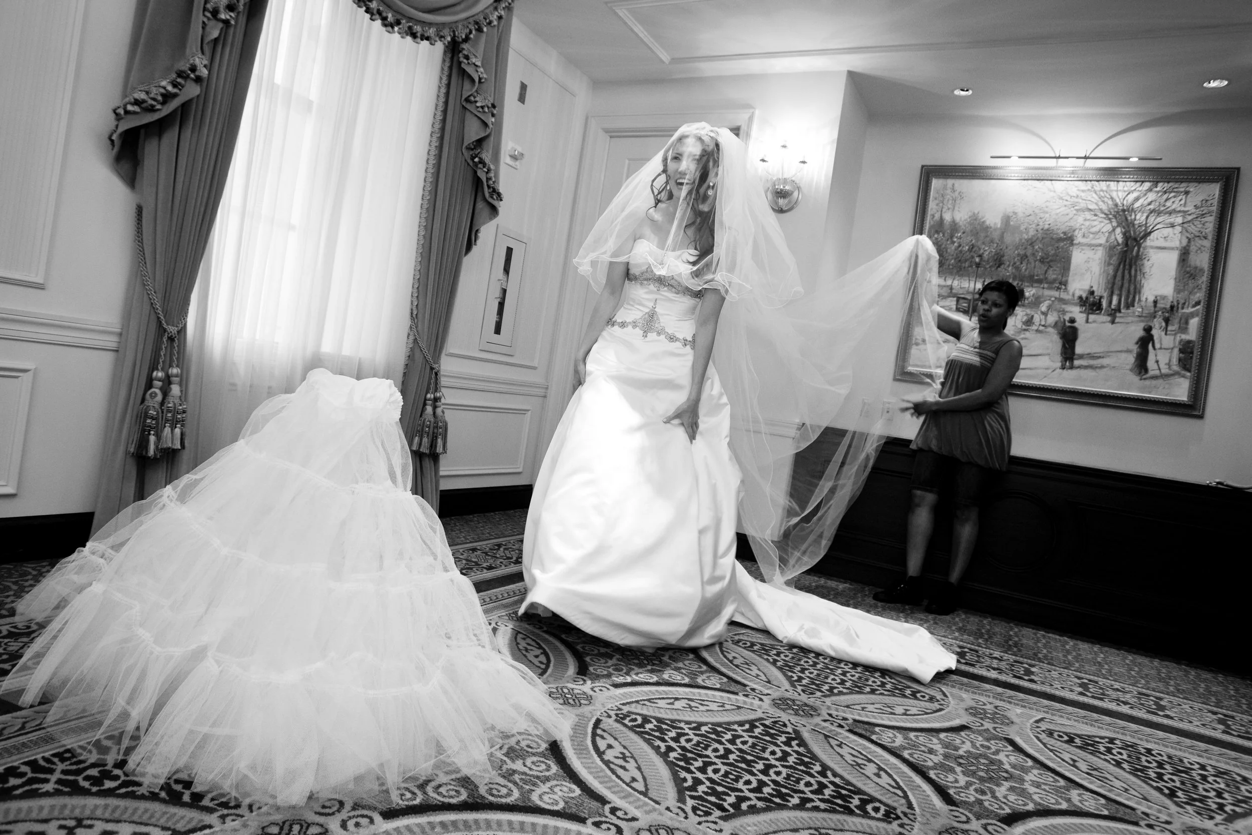 A bride in a wedding dress, with a veil, is smiling as a woman helps her with her gown in a decorated hotel room.