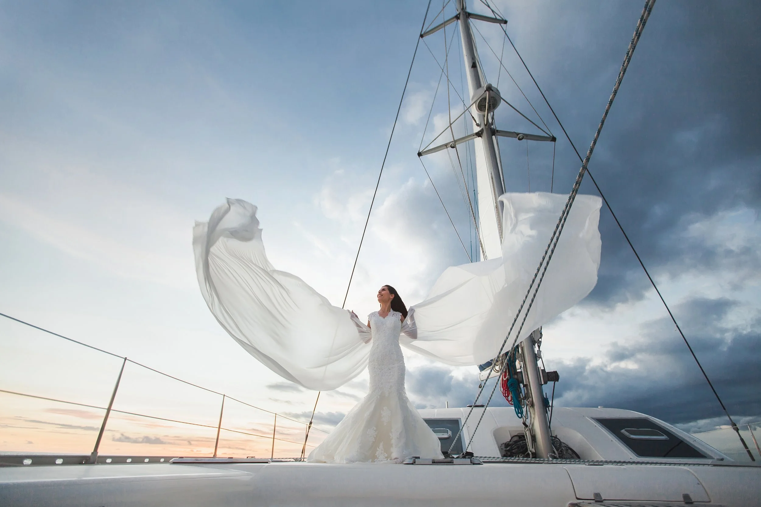 A woman in a white wedding dress standing on a sailboat at sunset, holding up flowing fabric that looks like wings, with a cloudy sky in the background.