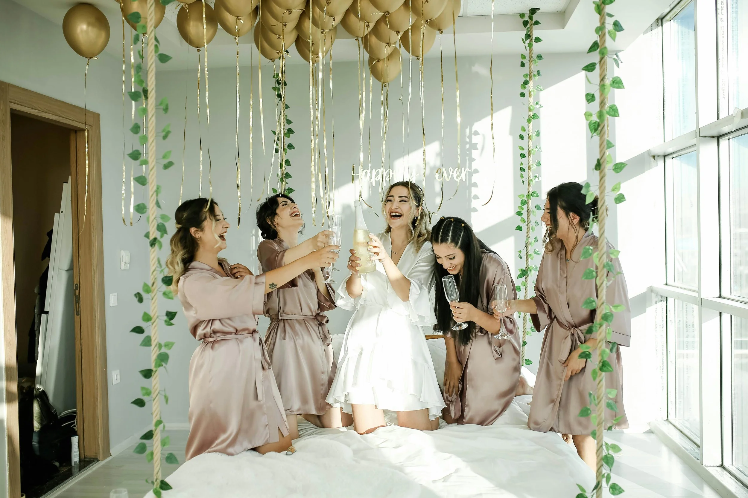 A group of women celebrating in a bright room decorated with balloons and hanging greenery, wearing satin robes, with champagne and glasses, laughing and enjoying a joyful moment, possibly at a bridal shower or bachelorette party.