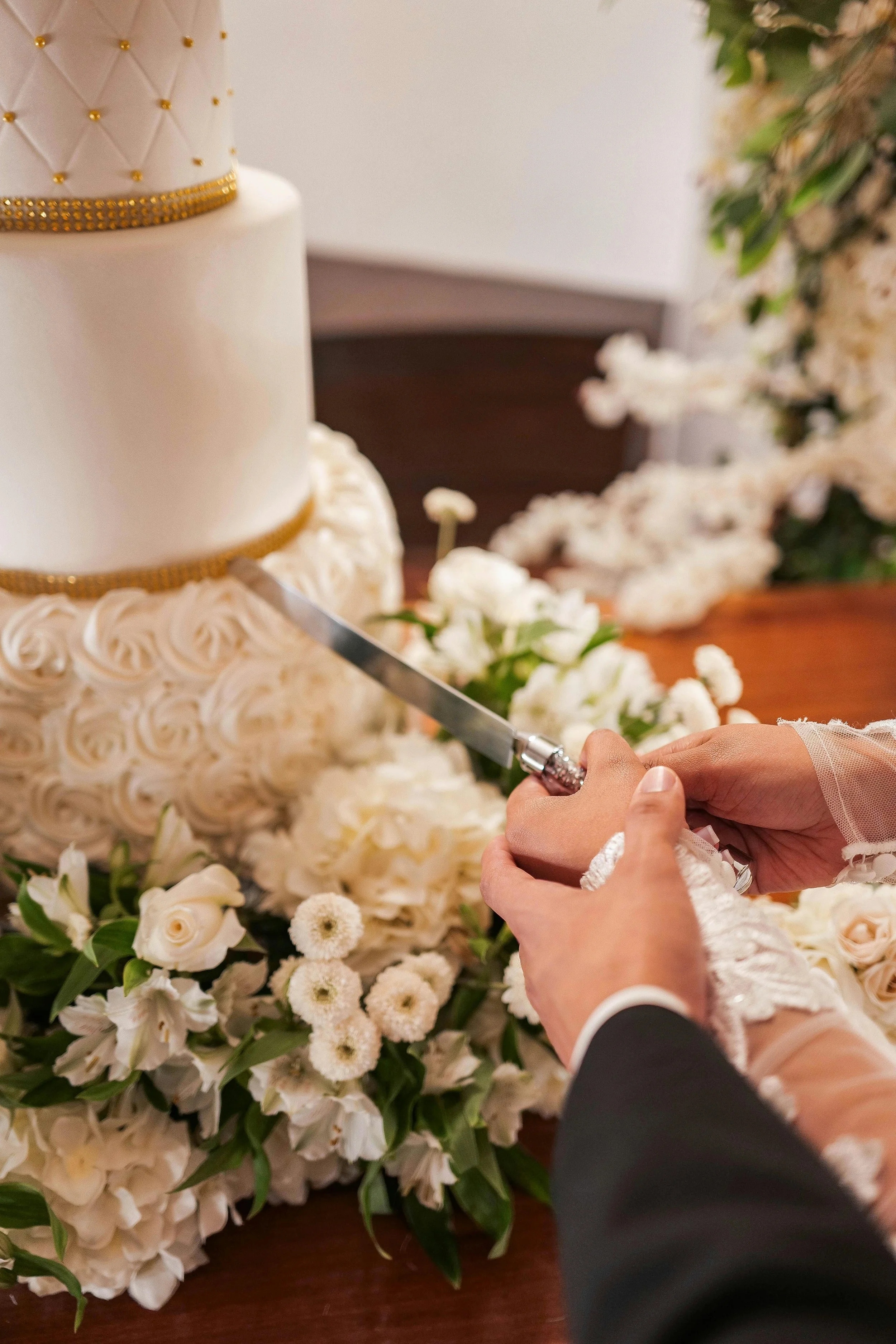 Close-up of a person cutting a wedding cake with a knife, surrounded by white flowers, at a wedding celebration.