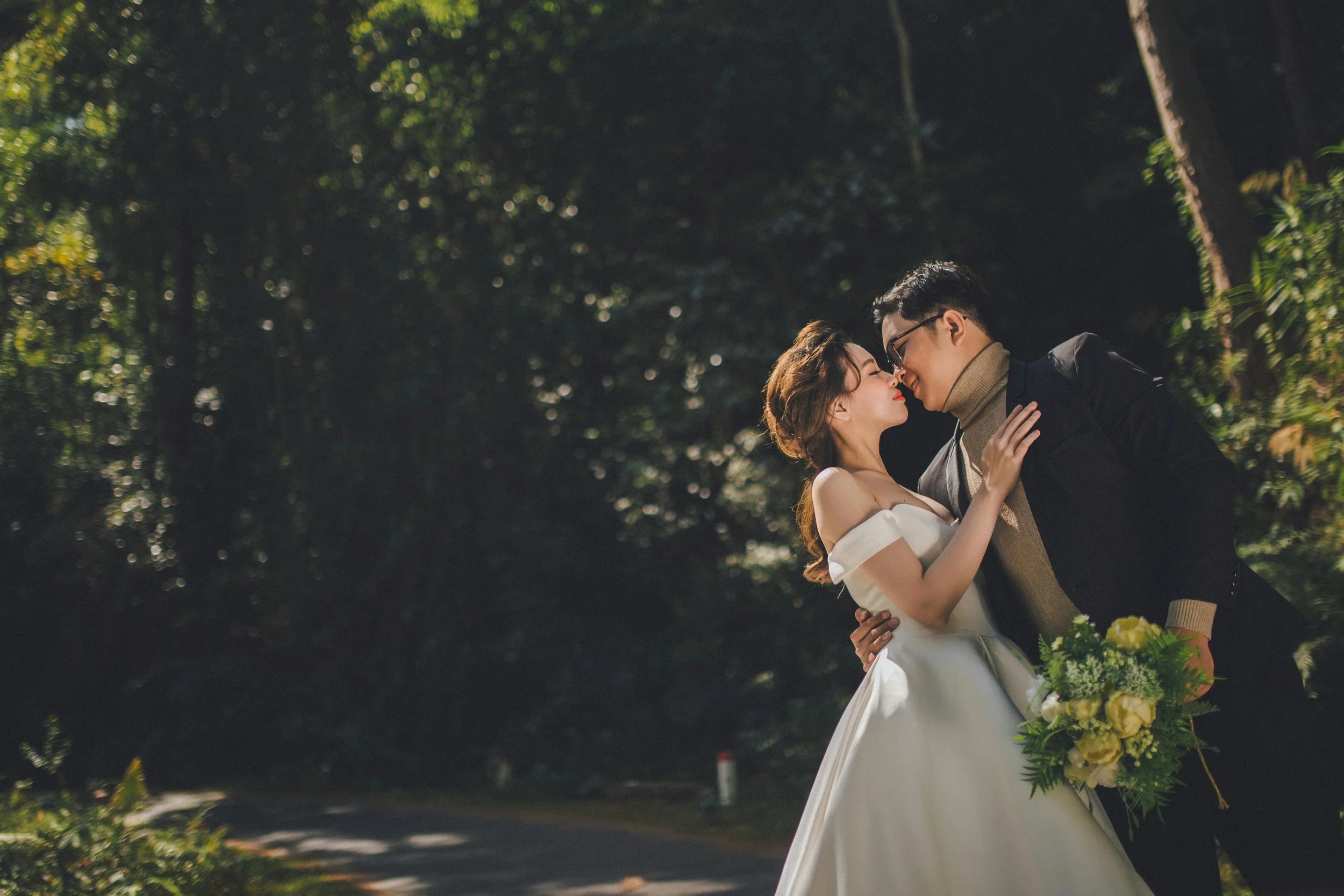 A bride and groom kissing outside in a wooded area, with the bride holding a bouquet of yellow roses.