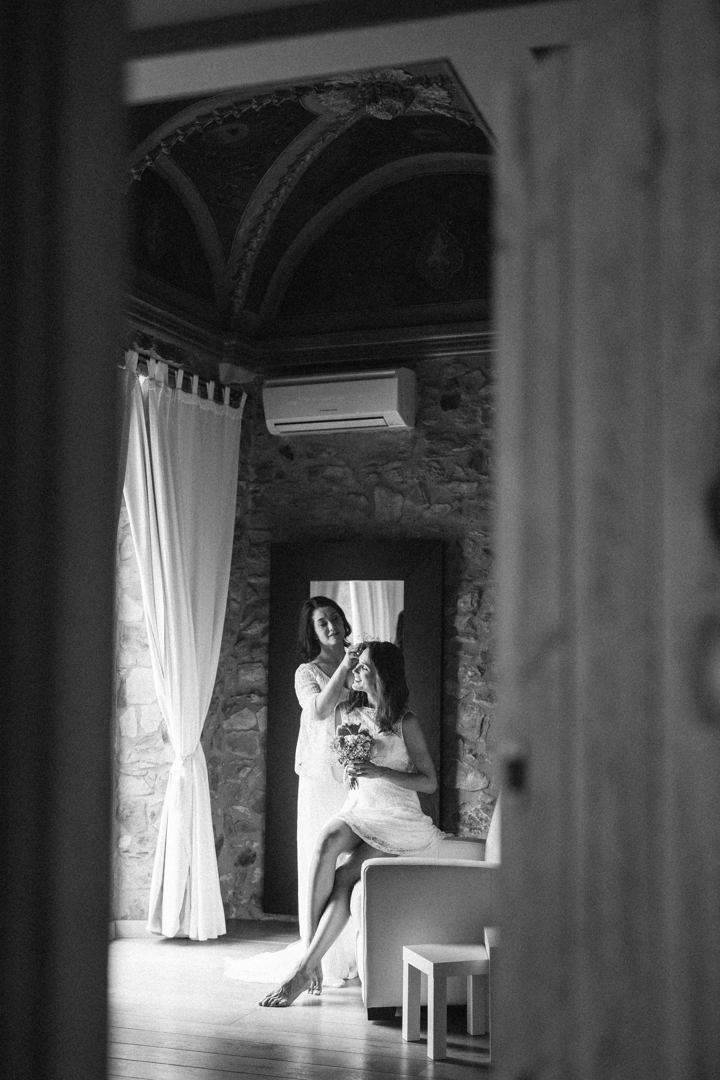 A black-and-white photo of two women, one sitting on a chair holding a bouquet, and a second woman standing and brushing the seated woman's hair, reflected in a mirror, in a room with stone walls and a curtain.