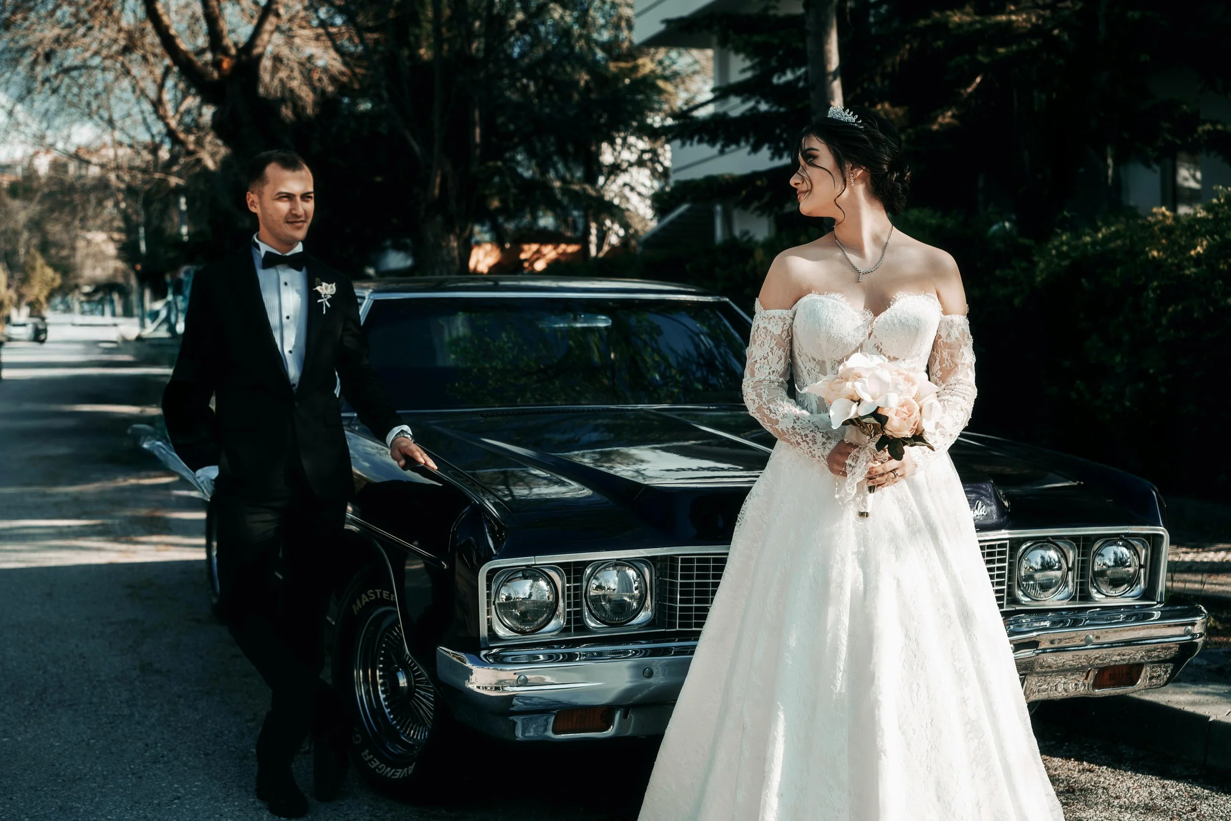 Bride in white wedding dress holding bouquet looking at groom in black tuxedo standing near a classic black car on a tree-lined street.