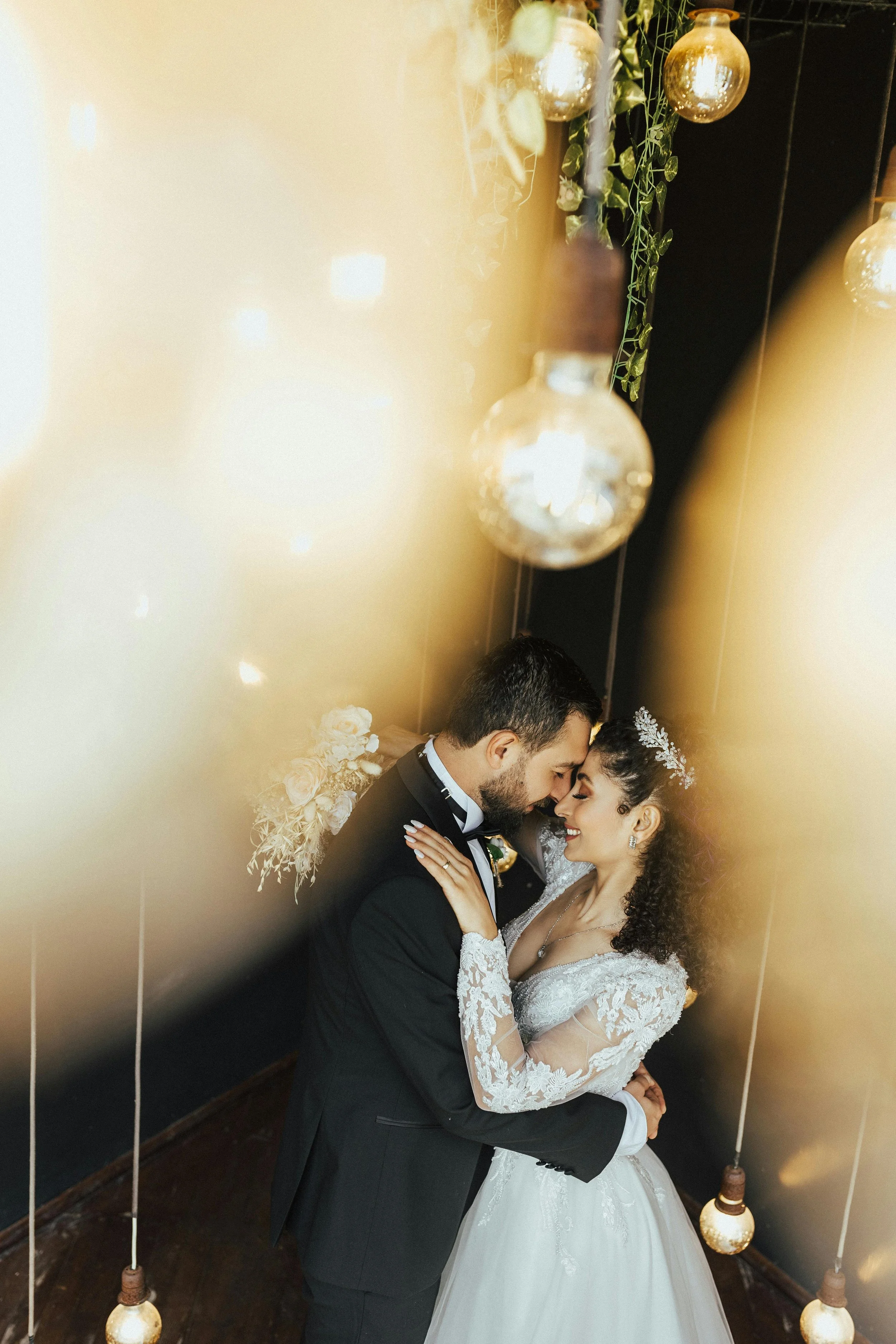 A bride and groom embracing each other at their wedding, with decorative hanging lights and floral decorations around them.