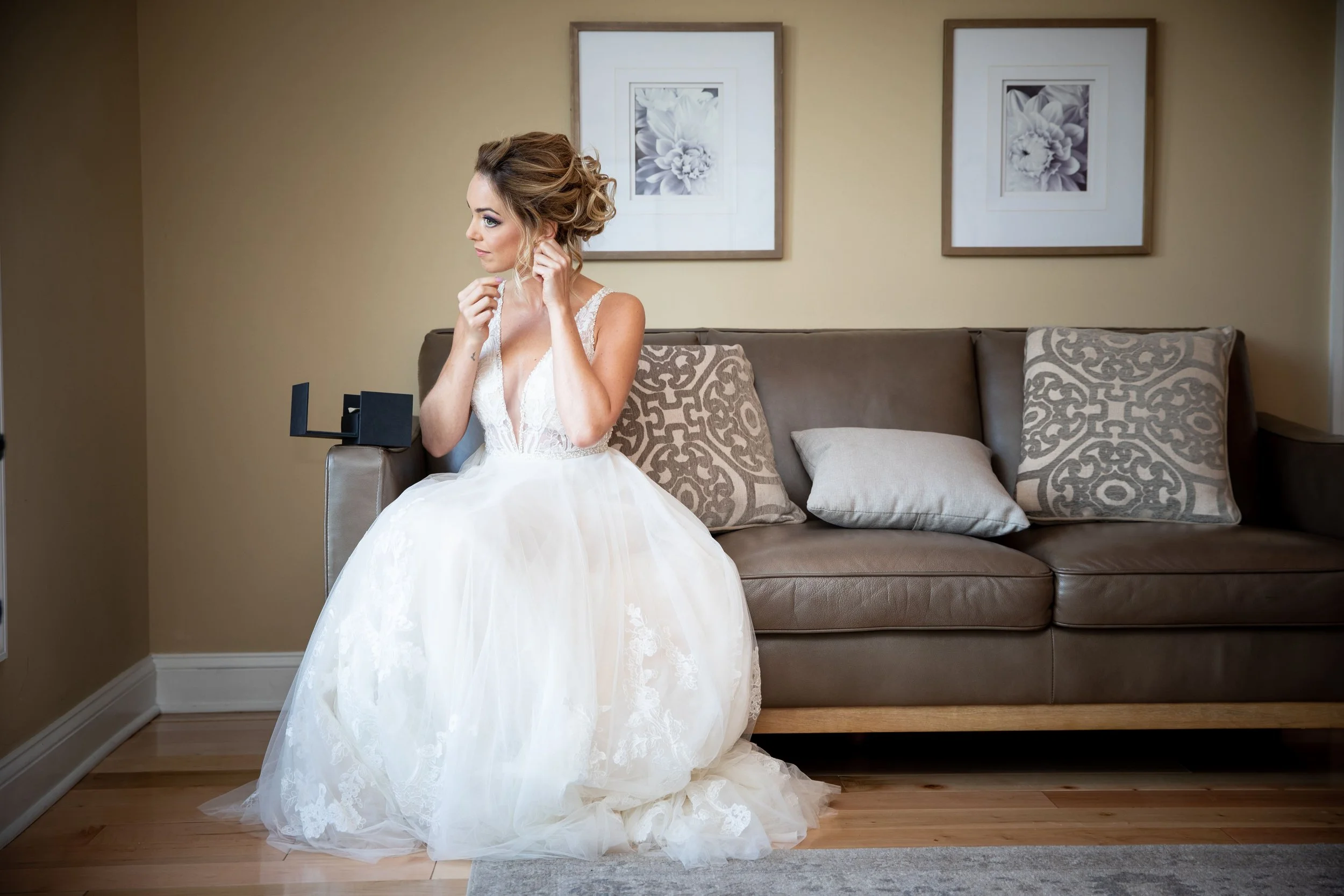 A bride in a white wedding dress sitting on a sofa, adjusting her earring, with two framed floral pictures on the wall behind her.