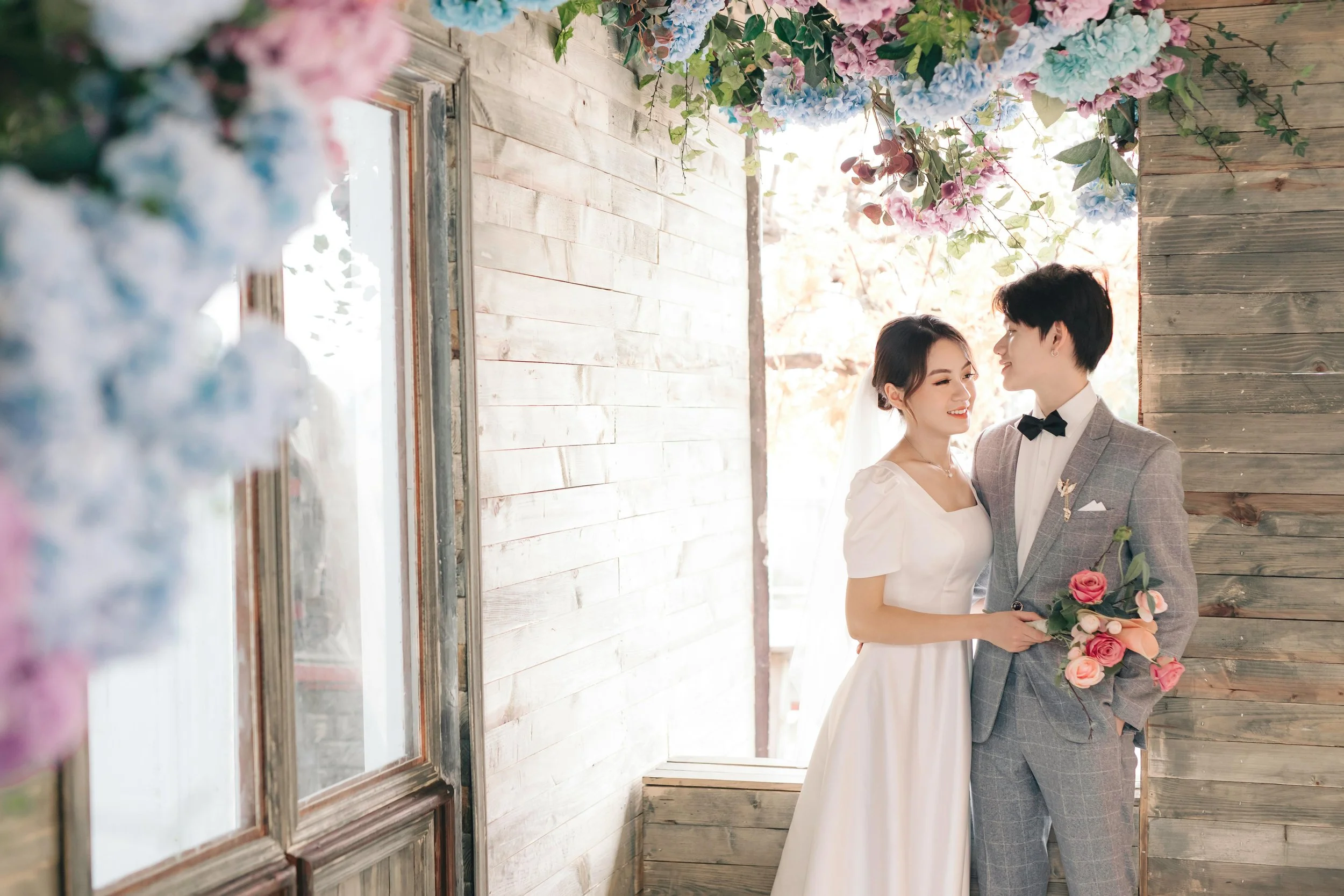 A bride and groom stand close together in a wedding setting, surrounded by wooden walls and floral decorations, smiling at each other. The bride holds a bouquet, and the groom is dressed in a gray checkered suit with a bow tie.