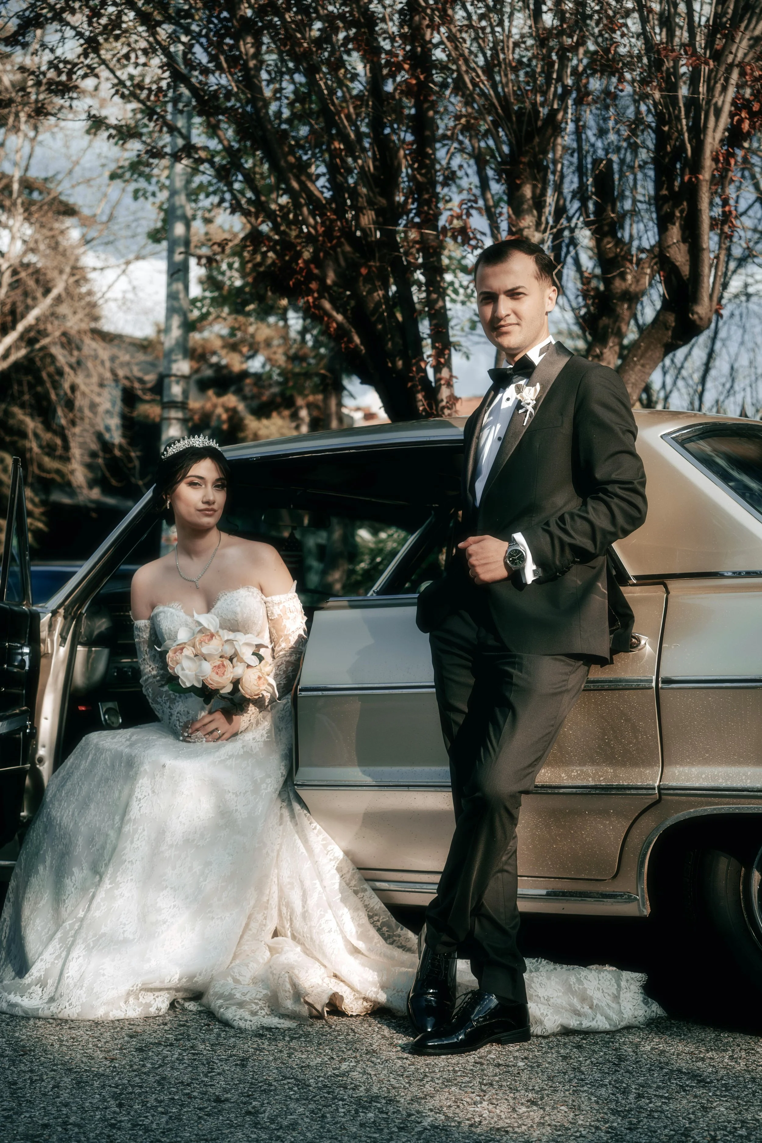 A bride in a white wedding dress holding a bouquet of flowers sitting beside a groom in a black tuxedo standing next to a vintage car outdoors on a sunny day.