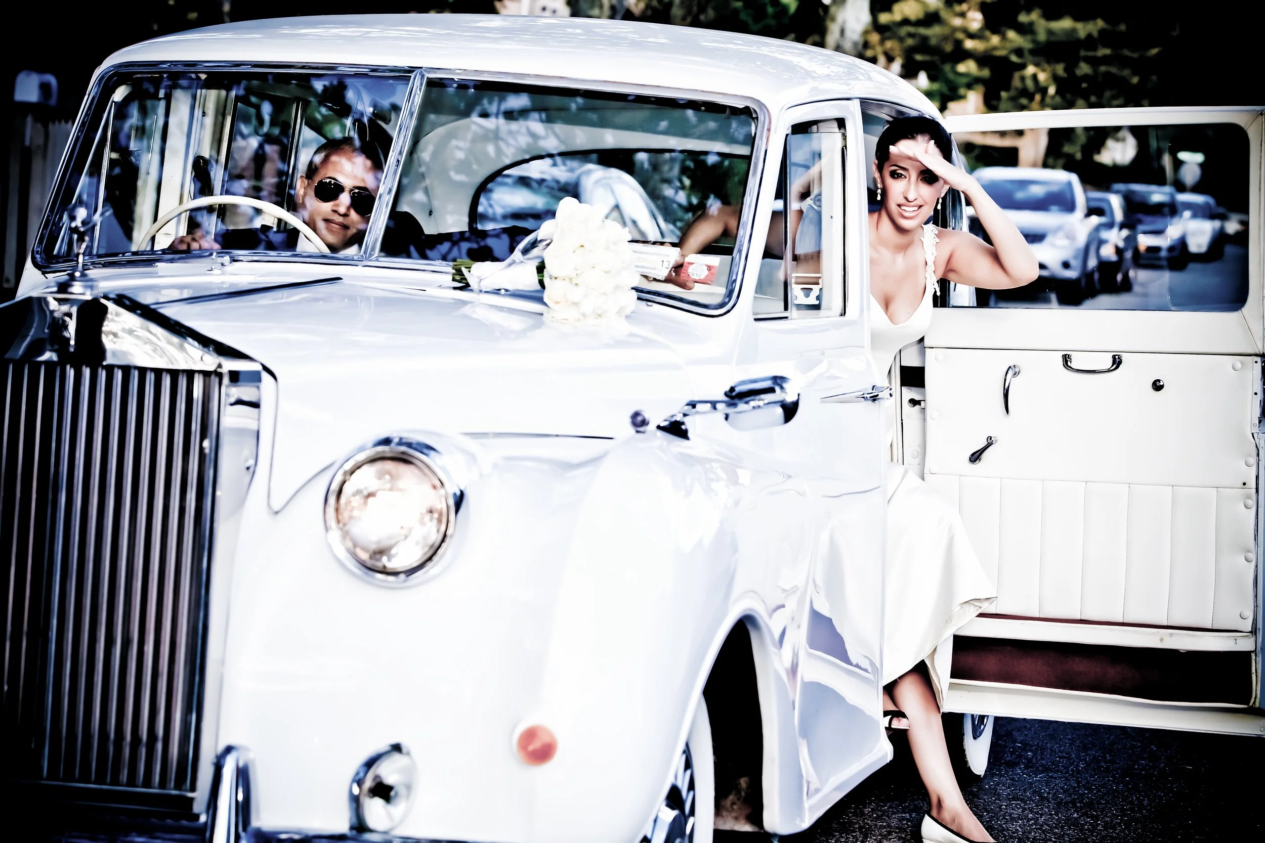 A bride in a white wedding dress sitting in an open door of a vintage white car with a bouquet on the dashboard, waving and smiling, with a person wearing sunglasses driving, and other parked vehicles in the background.