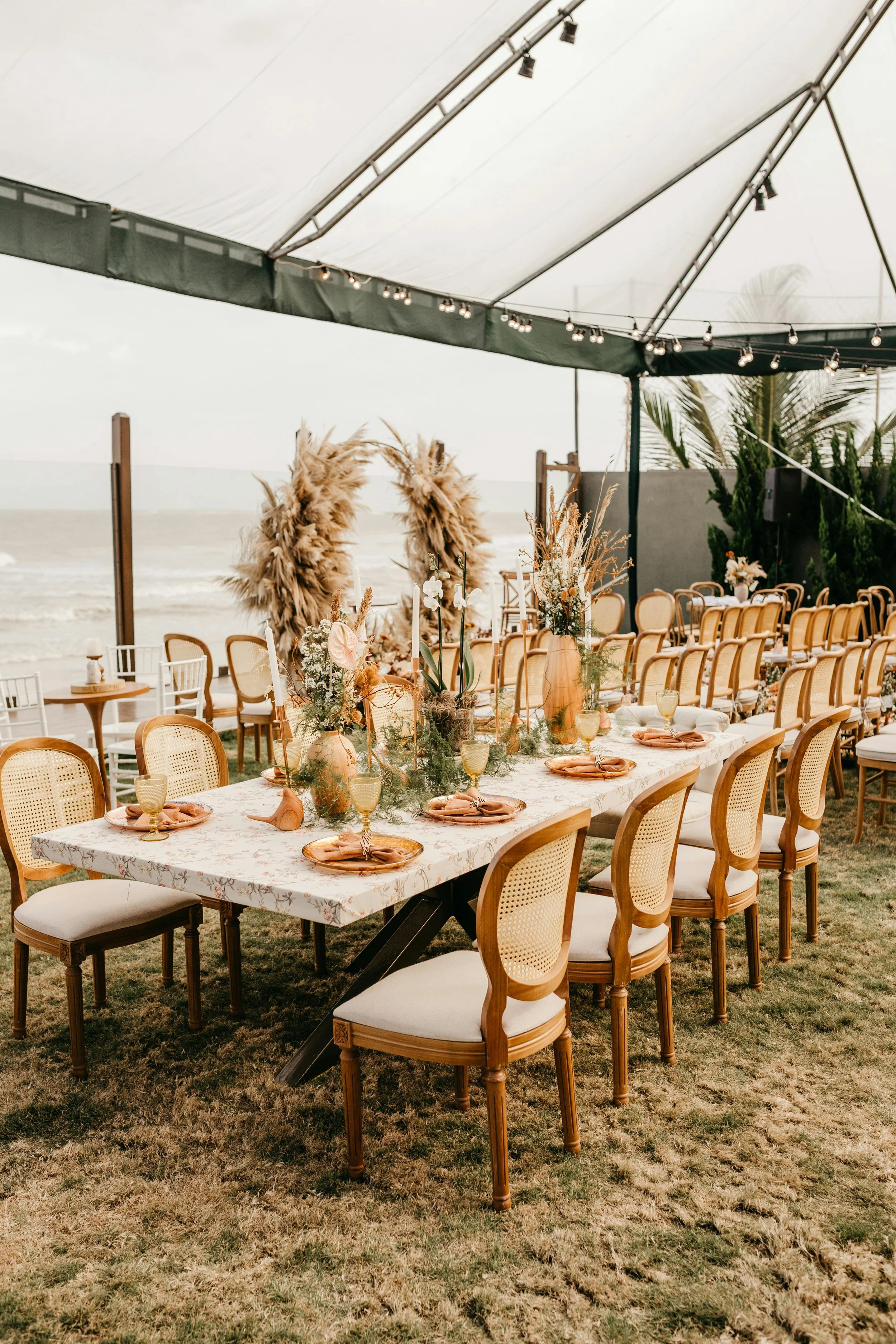 Beachside wedding reception setup with a long floral table, surrounded by chairs, under a tent, with decorative pampas grass and ocean in the background.