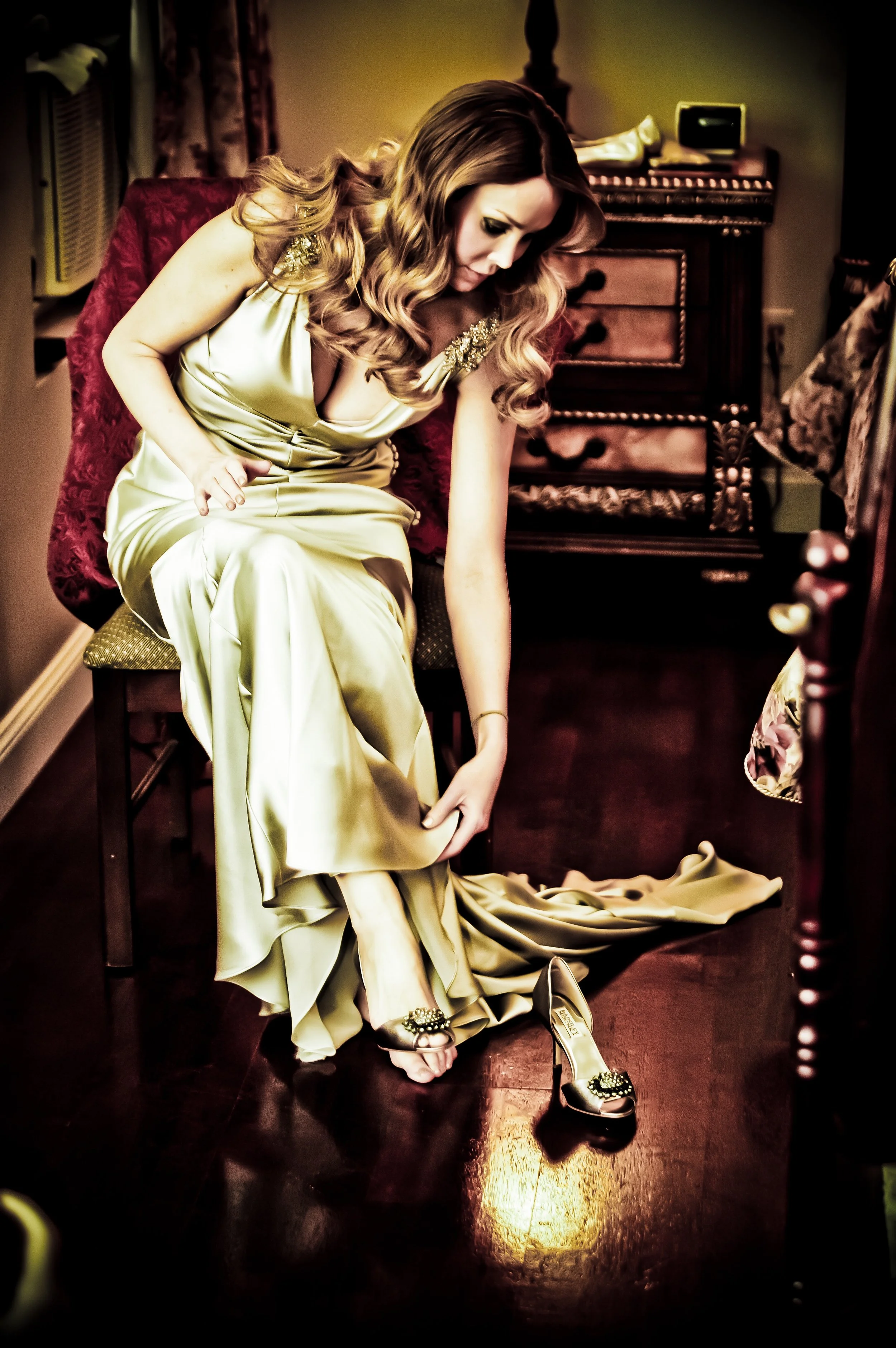 A woman sitting on a red velvet chair, wearing a gold satin gown, is removing her shoe as she tries to put on her other shoe on a polished wooden floor. The room has vintage furniture and warm lighting.