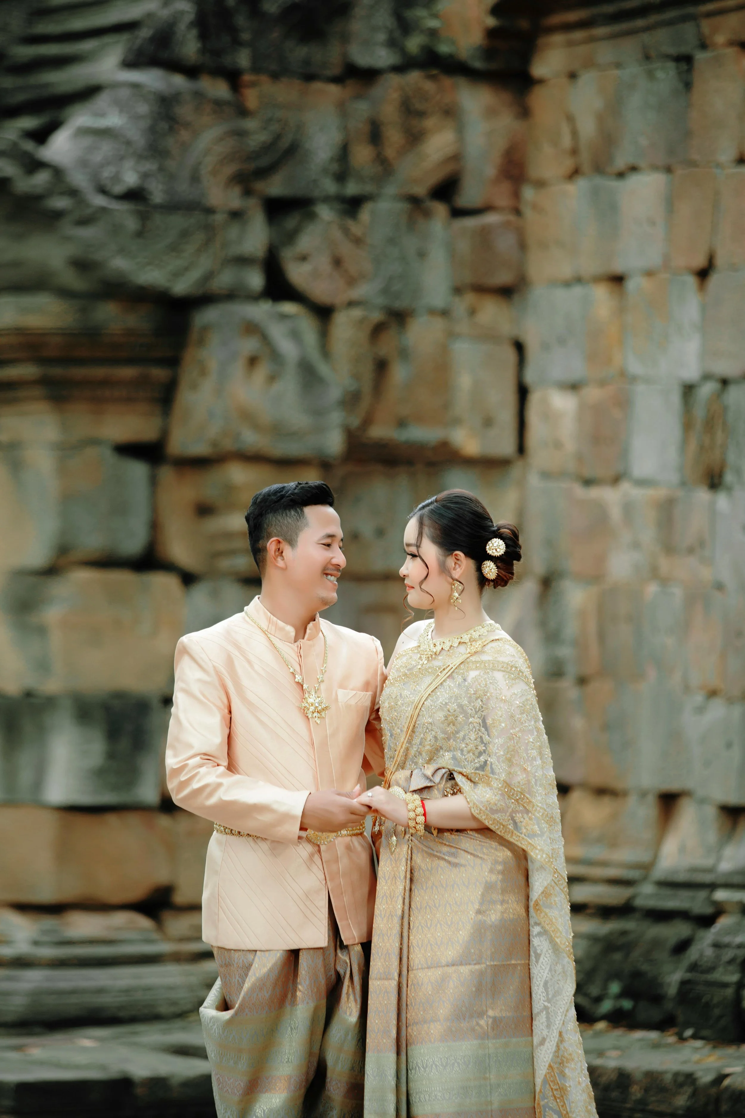 A couple dressed in traditional Thai wedding attire standing close together in front of an ancient stone wall, holding hands and smiling at each other.