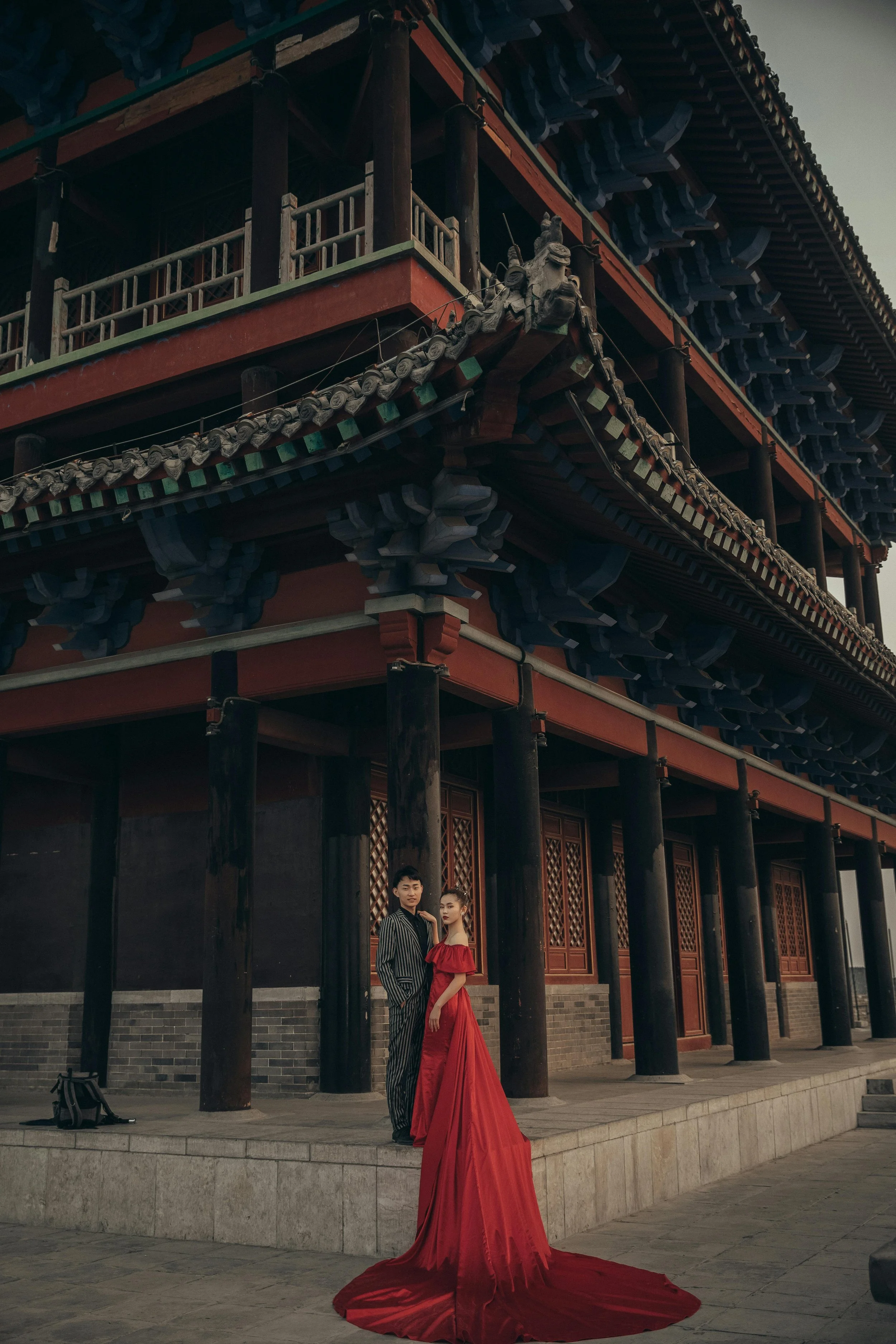 A young man and woman in formal attire, the woman in a long red gown with a train, standing on a stone platform in front of a traditional multi-story Asian temple with ornate wooden architecture.