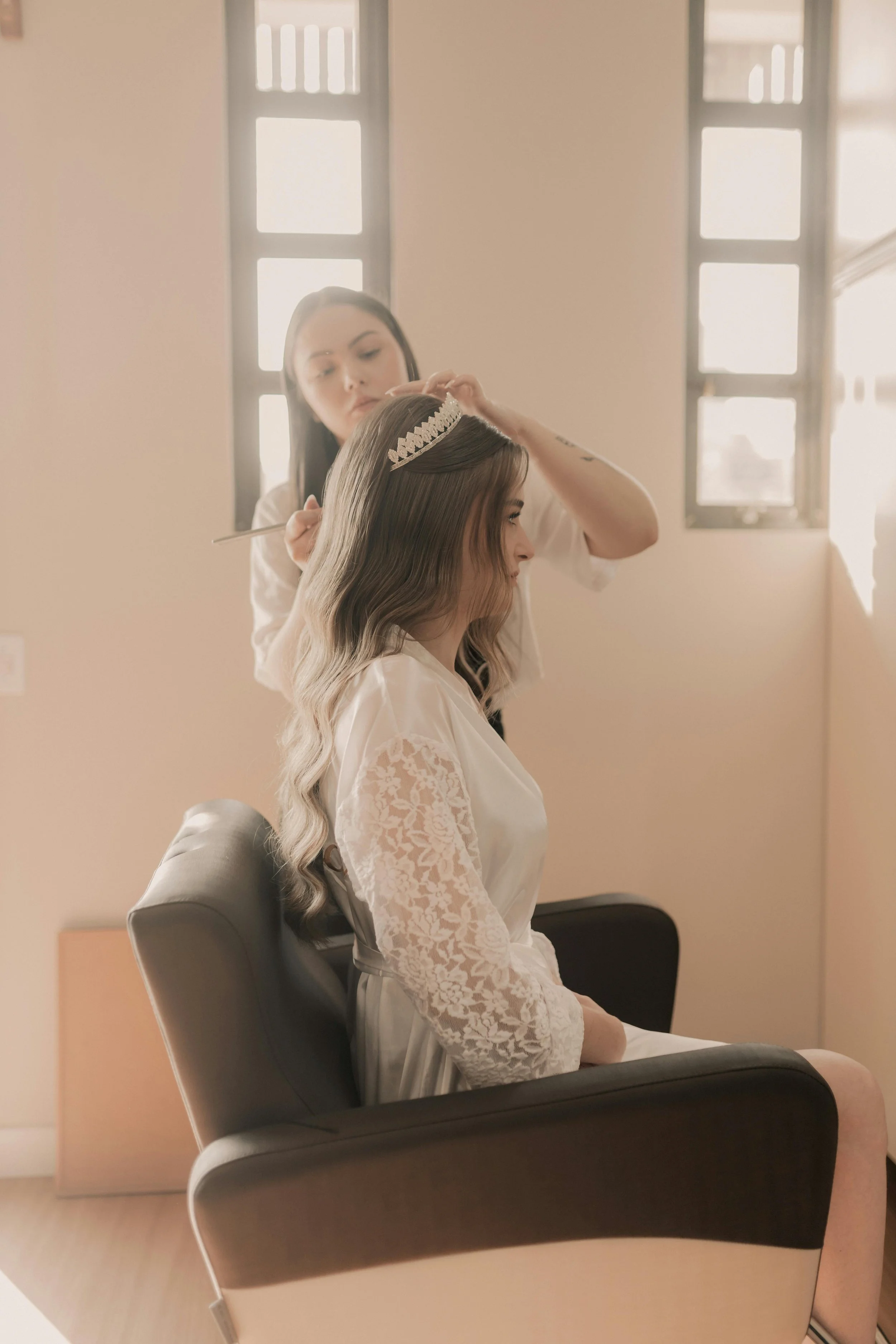 A woman with long, wavy hair is sitting in a chair while another woman styles her hair and places a tiara on her head in a well-lit room with windows.