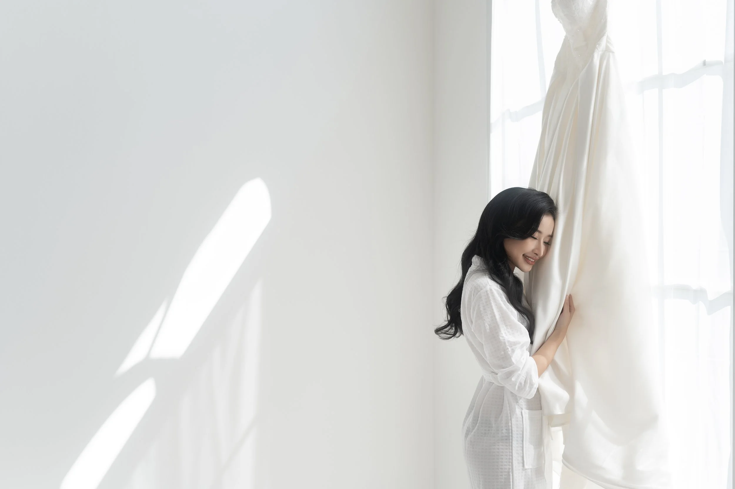 A woman with long black hair smiling and looking down, standing next to a large window with sunlight streaming in, holding a white curtain.