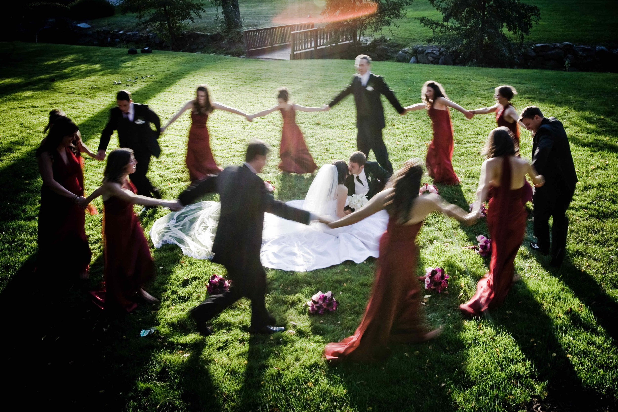 A wedding celebration in a grassy outdoor area with the bride and groom sitting on the ground surrounded by guests, forming a circle and holding hands. The guests are dressed in formal attire, with women in red dresses and men in black suits. The sce
